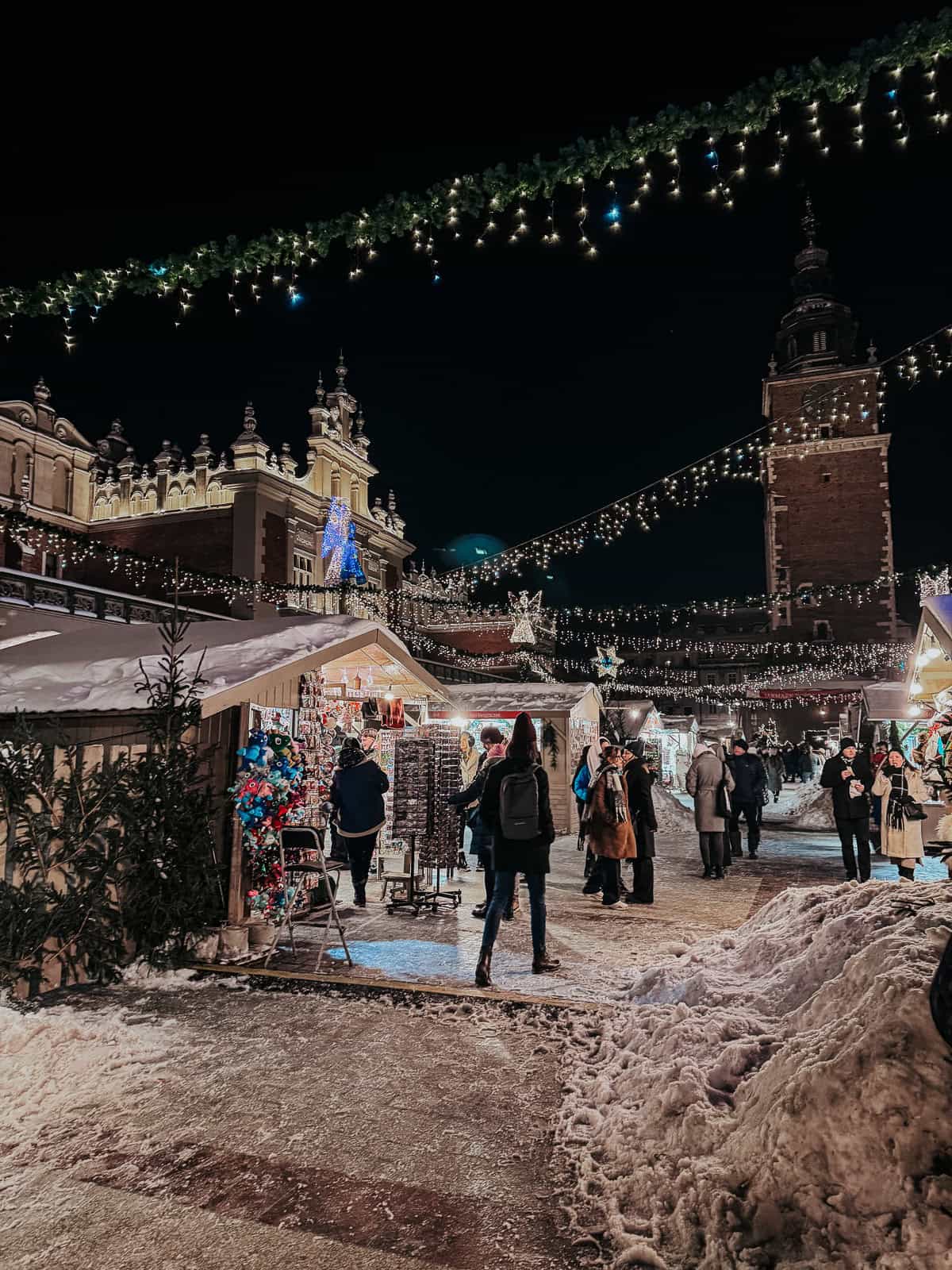 Night scene at a Christmas market with a view of the historic cloth hall illuminated by festive lights, snowy paths, and shoppers browsing through various stalls