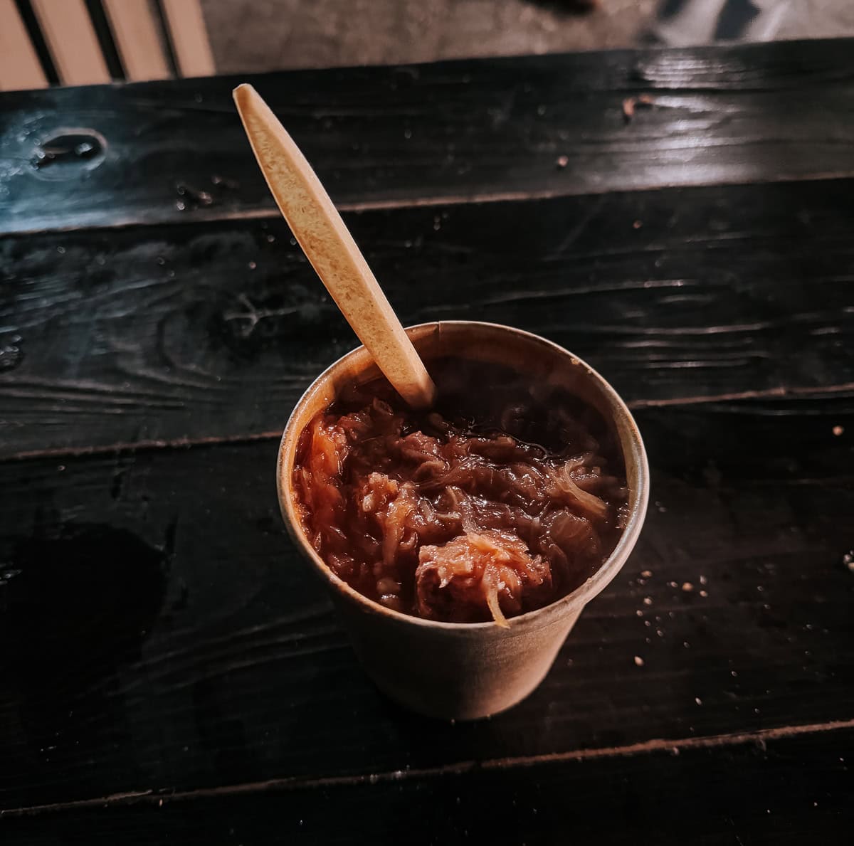 A paper cup of steaming bigos (traditional Polish stew of cabbage and meat) on a dark wooden table, with a wooden spoon, captured in an intimate, moody lighting
