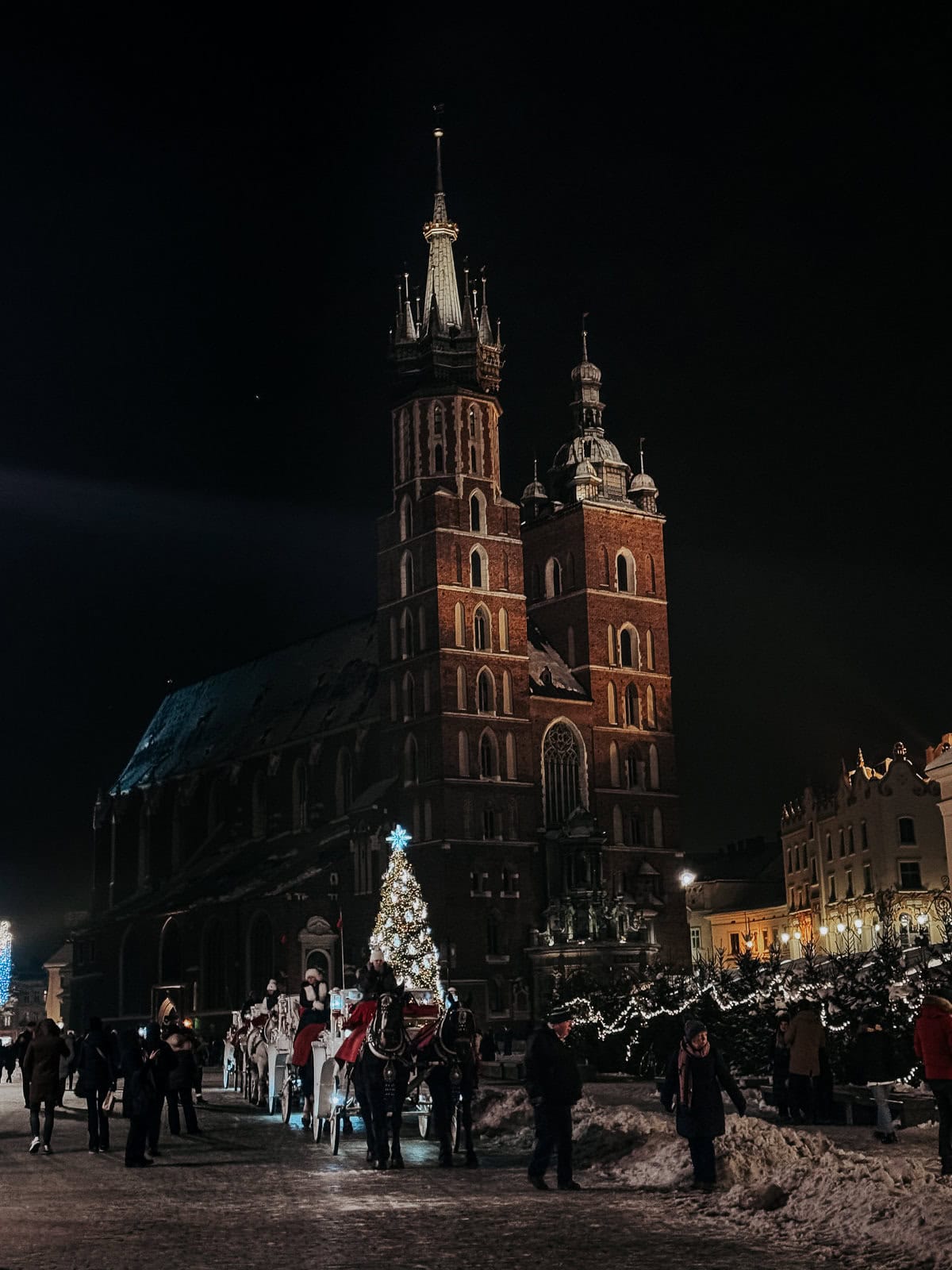 A captivating night scene of St. Mary's Basilica in Krakow, dramatically illuminated against the dark sky. The scene includes a beautifully lit Christmas tree and a horse-drawn carriage, surrounded by people enjoying the festive atmosphere at the Christmas market