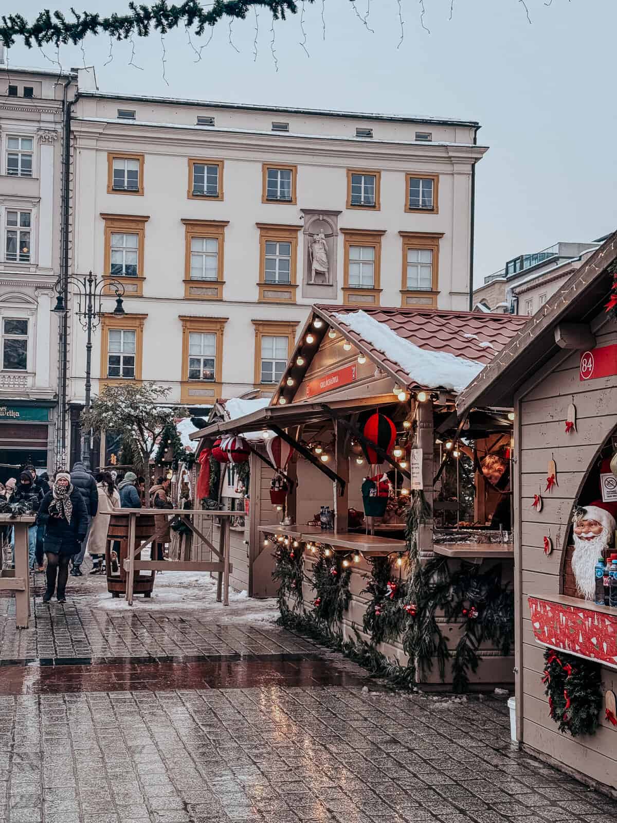 Festive wooden stall richly decorated with Christmas ornaments and lights, offering holiday delicacies under a snowy awning, with busy shoppers in winter attire at a European Christmas market.