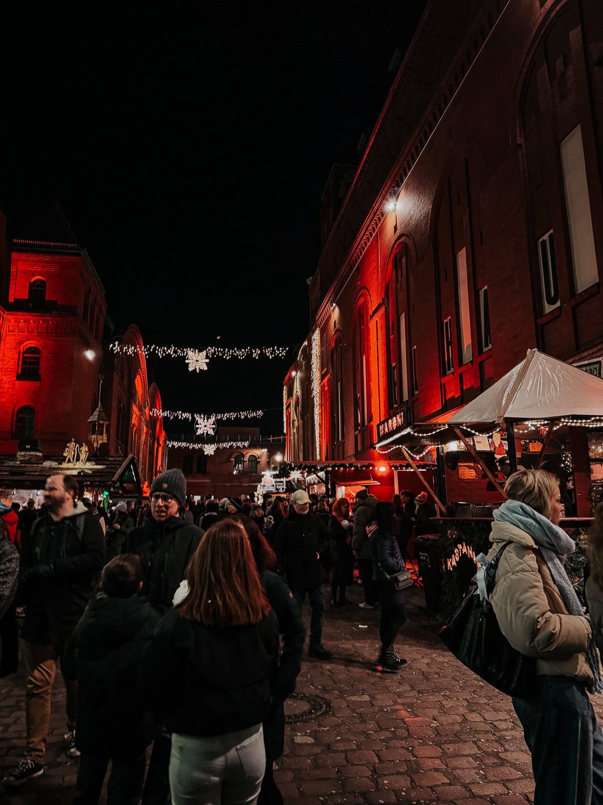 A vibrant evening scene at a holiday market, with bustling crowds of people in winter clothing. The scene is illuminated by festive lights strung above and the warm glow of market stalls against a historic red brick building
