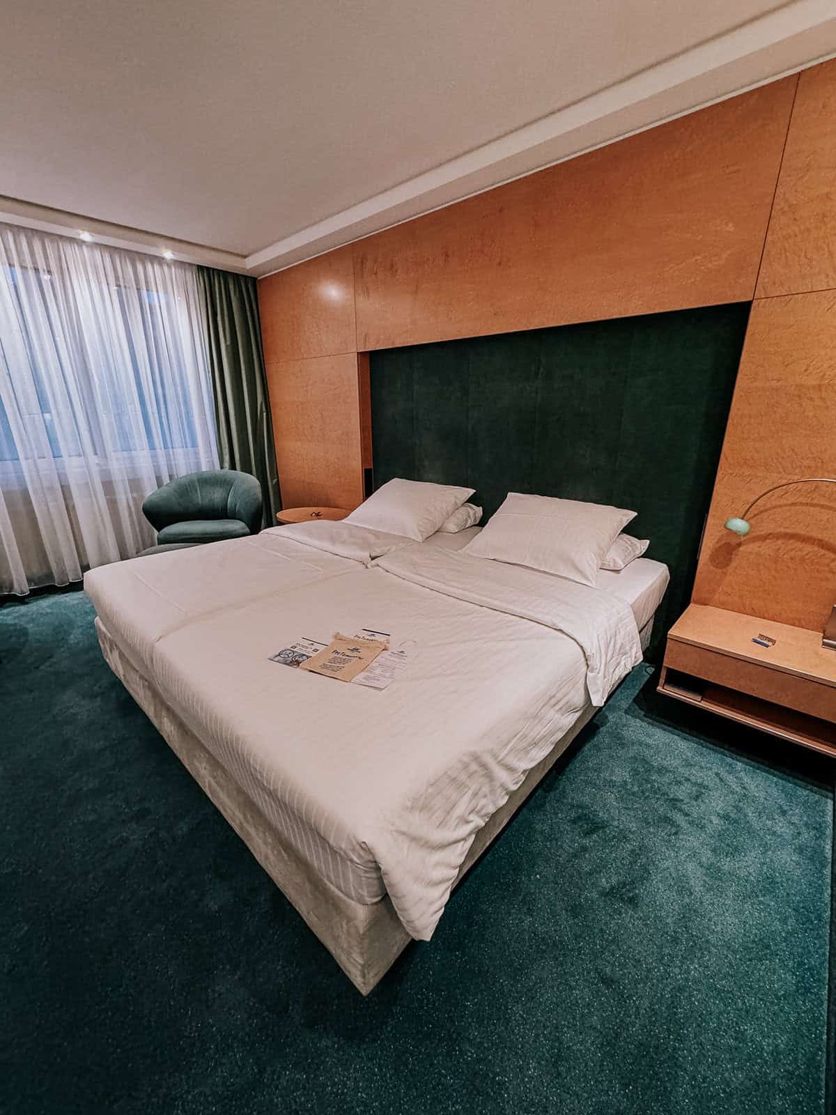 A spacious hotel room featuring a large bed with white linens, a green armchair, and a green carpet. The room has a modern yet warm ambiance, with a wide window draped with sheer curtains.