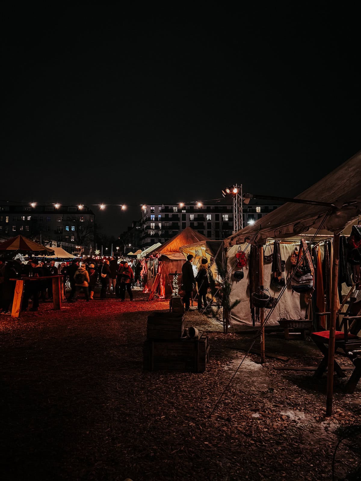 A night-time view of a medieval-themed outdoor market lit by string lights, with tents and stalls displaying various goods. The atmosphere is lively with visitors exploring the market on a ground covered with wood chips