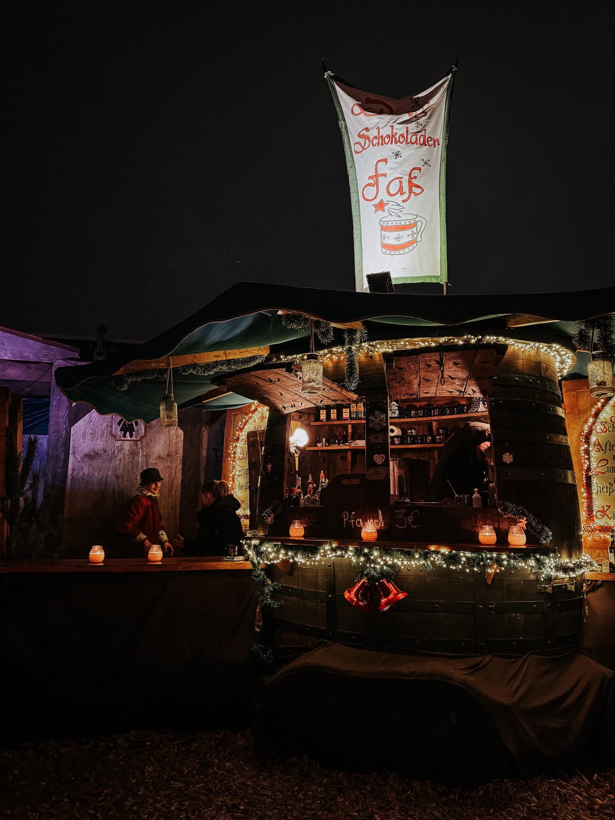An intimate night market scene featuring a booth styled like a giant barrel, selling hot beverages. The booth is festively decorated with Christmas lights and offers a warm ambiance in the evening chill.