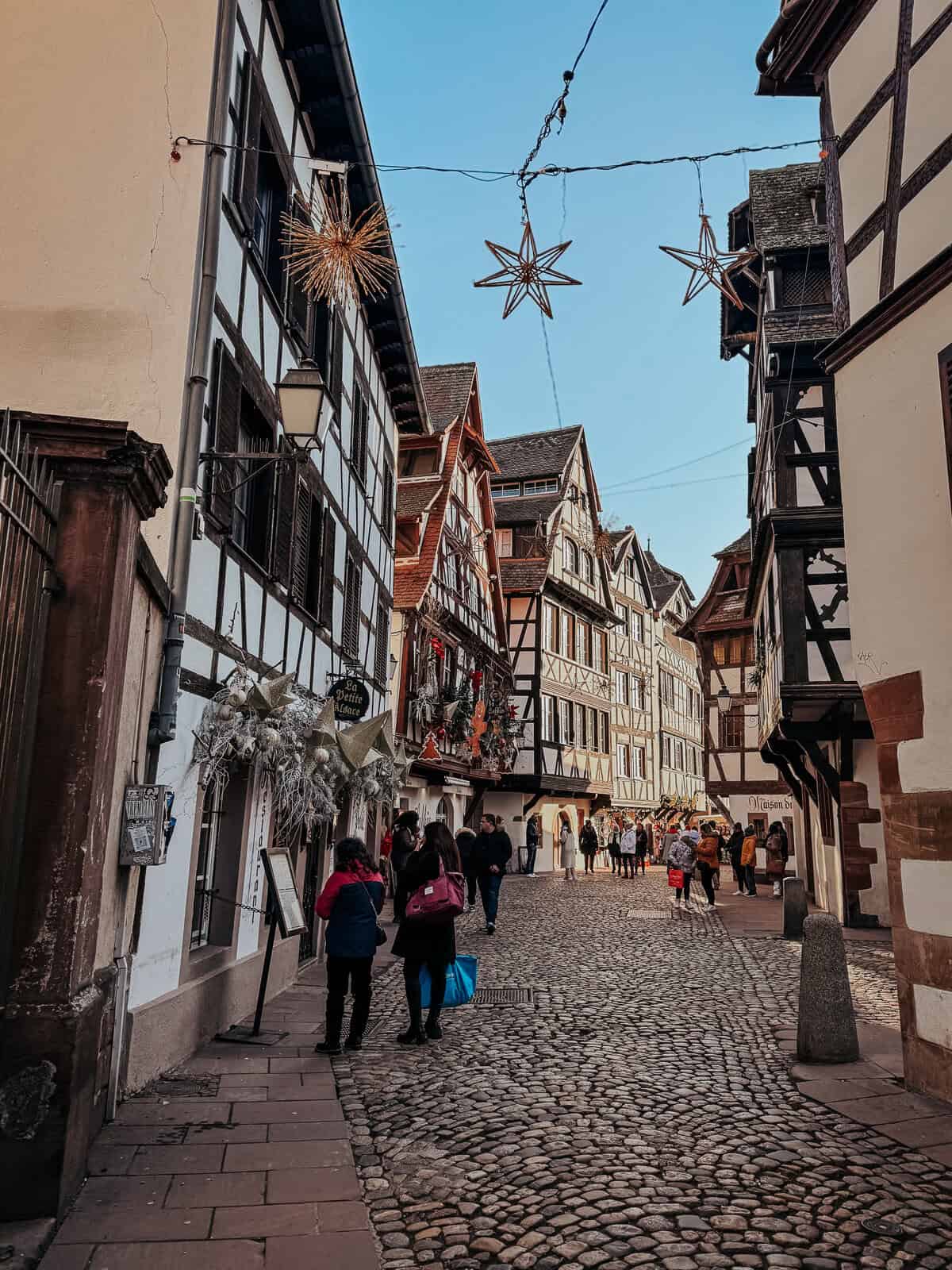 A bustling narrow street lined with traditional timber-framed houses and festive decorations, filled with shoppers enjoying the holiday atmosphere