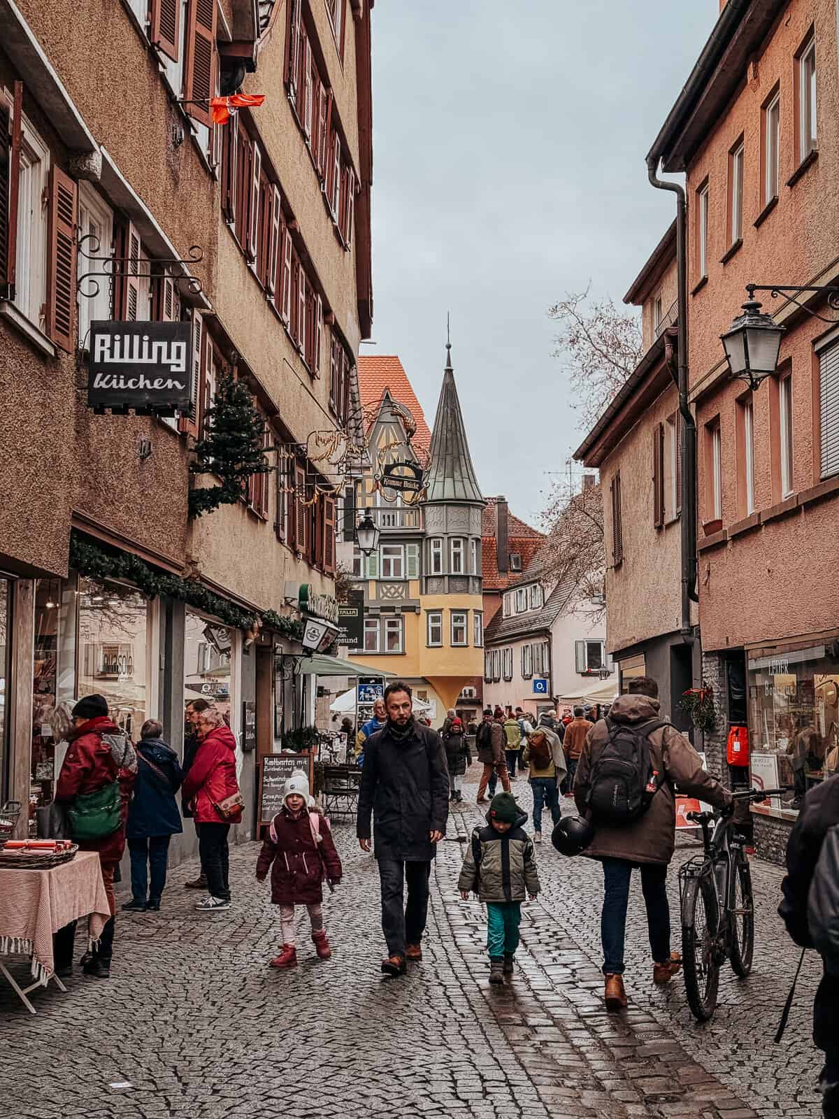 Residents and tourists walk through a cobblestone street in Tübingen, with colorful buildings, local shops like 'Rilling küchen', and festive street decorations visible