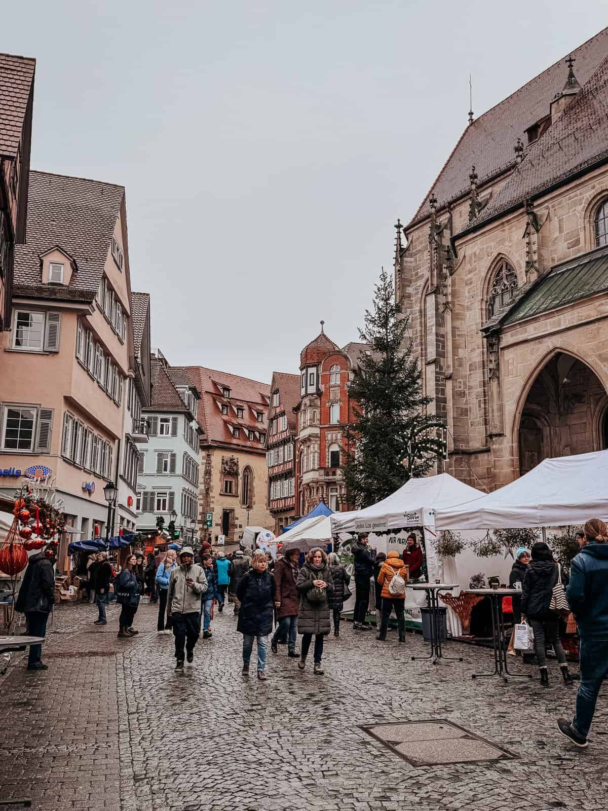 View of a busy street market adjacent to a large Gothic church in Tübingen, with people browsing stalls under overcast skies.