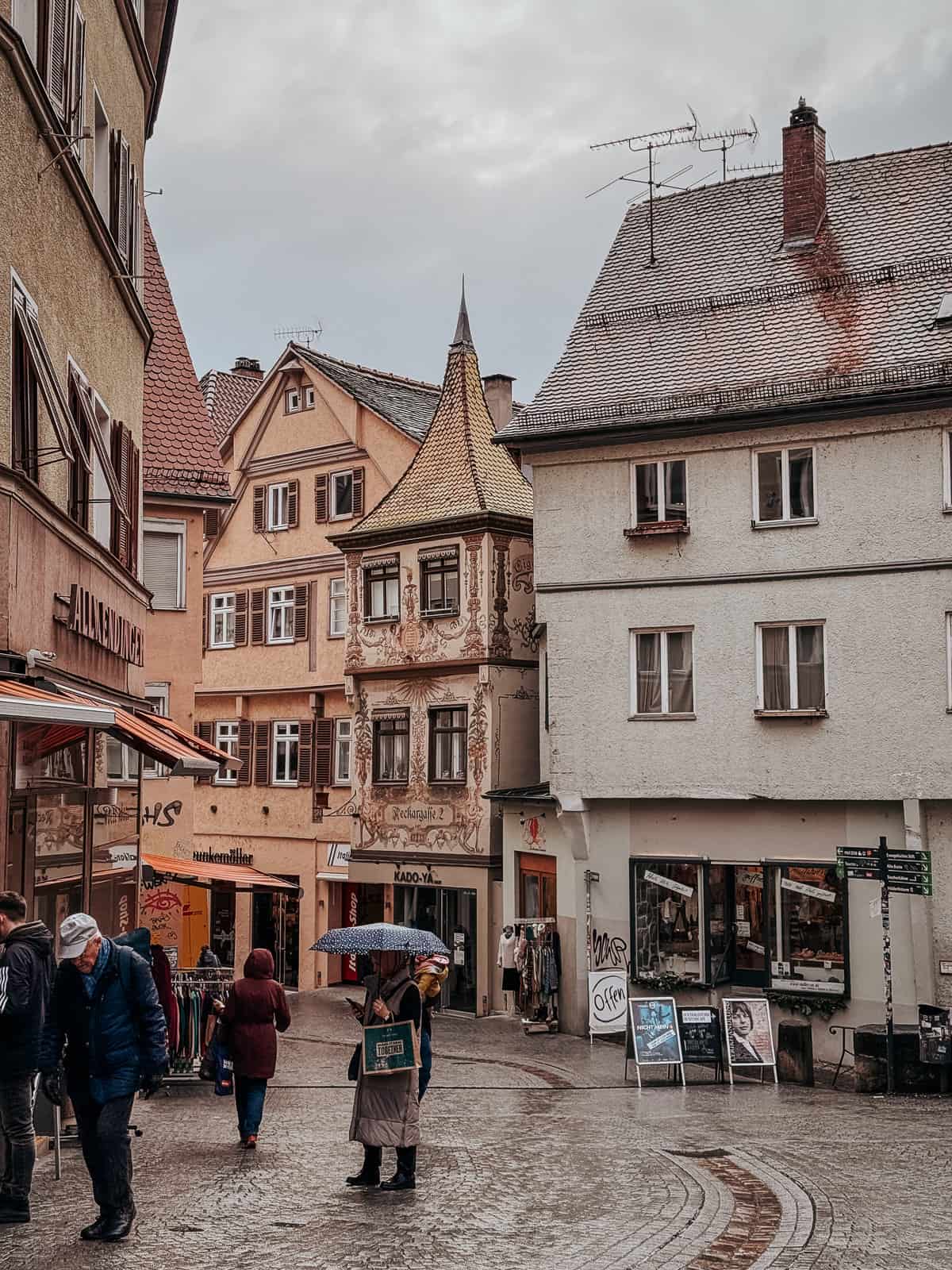 A cobblestone street in Tübingen, lined with people walking and beautifully detailed buildings, including one with ornate frescoed facades.