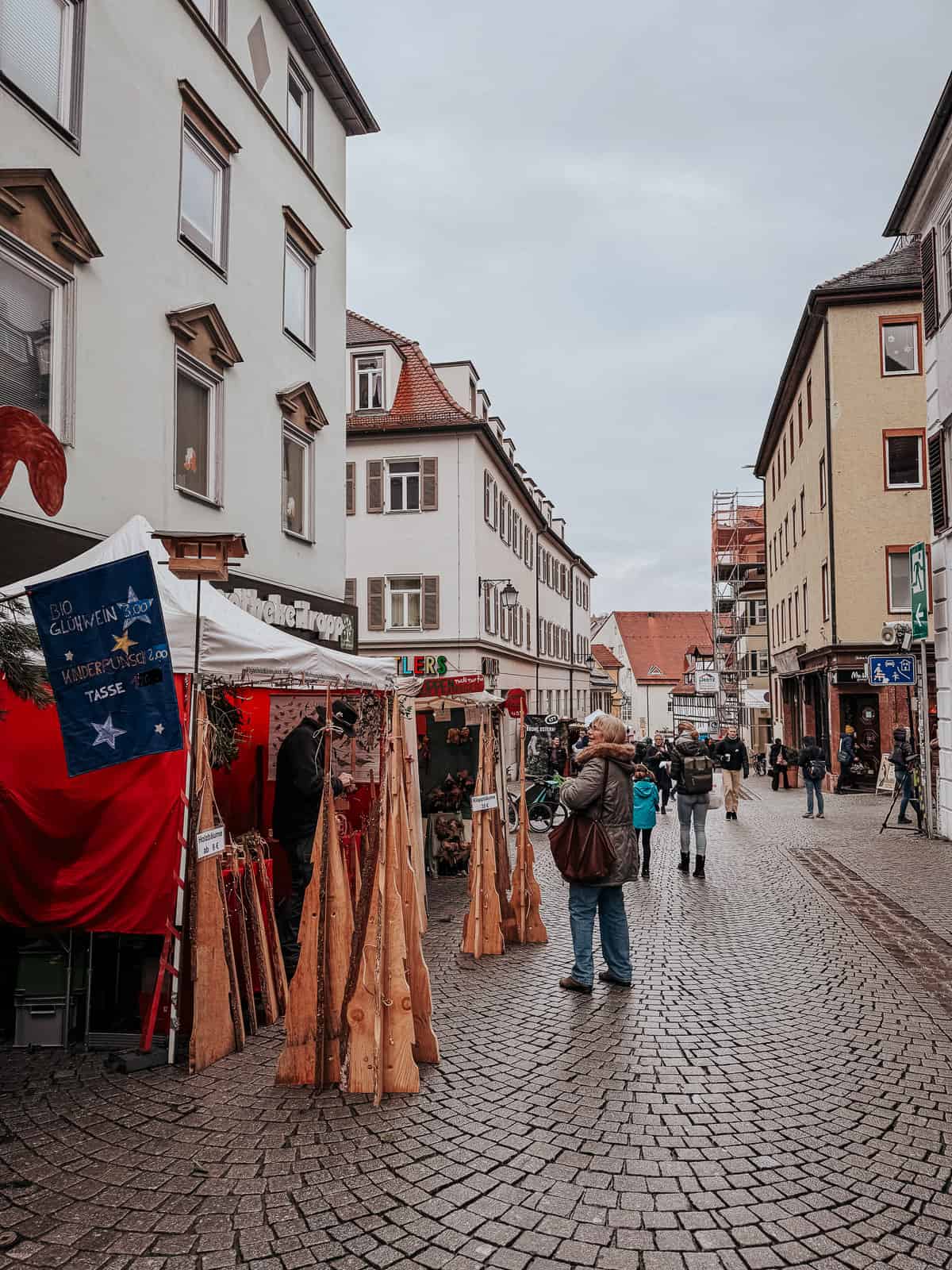 Cobblestone street in Tübingen bustling with people browsing items at various market stalls selling wooden artifacts and other handicrafts.