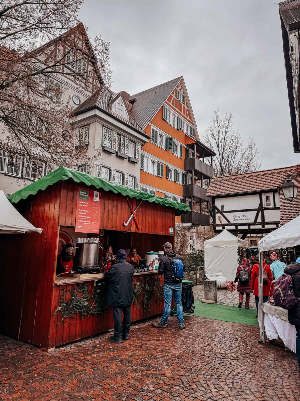 Outdoor winter market scene in Tübingen, showing a red wooden hut serving drinks and food, surrounded by people in winter attire