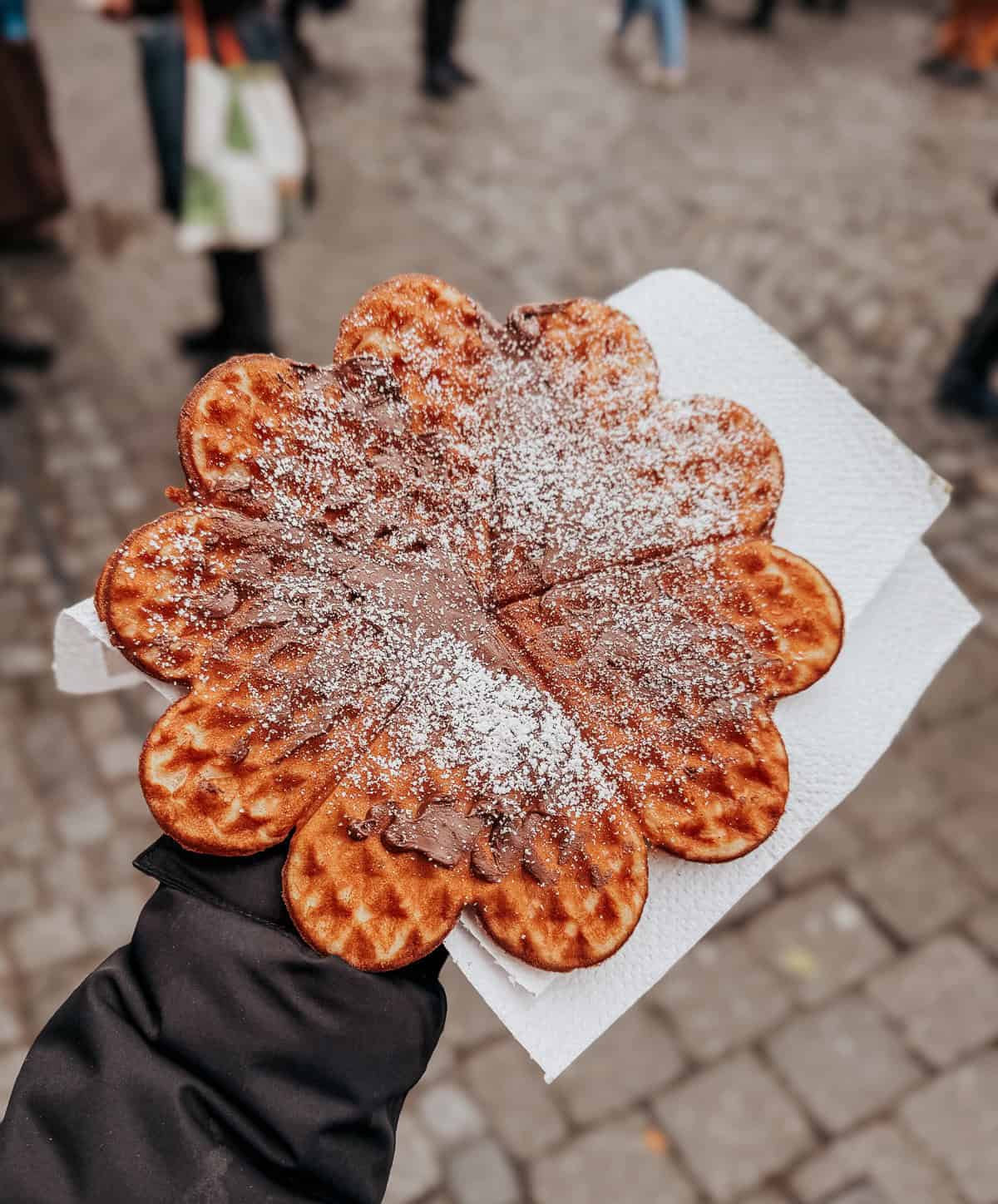 Close-up of a hand holding a traditional German pastry sprinkled with powdered sugar, with people in winter clothes blurred in the background.