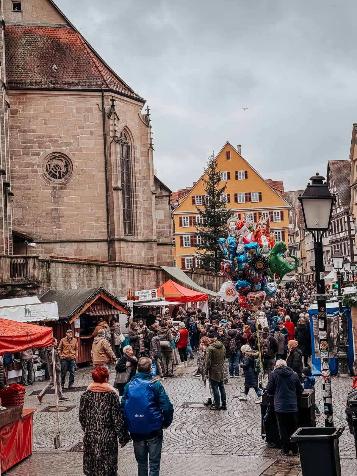 Crowded market square in Tübingen next to a large Gothic church, featuring vendors, shoppers, and colorful balloons, under an overcast sky.