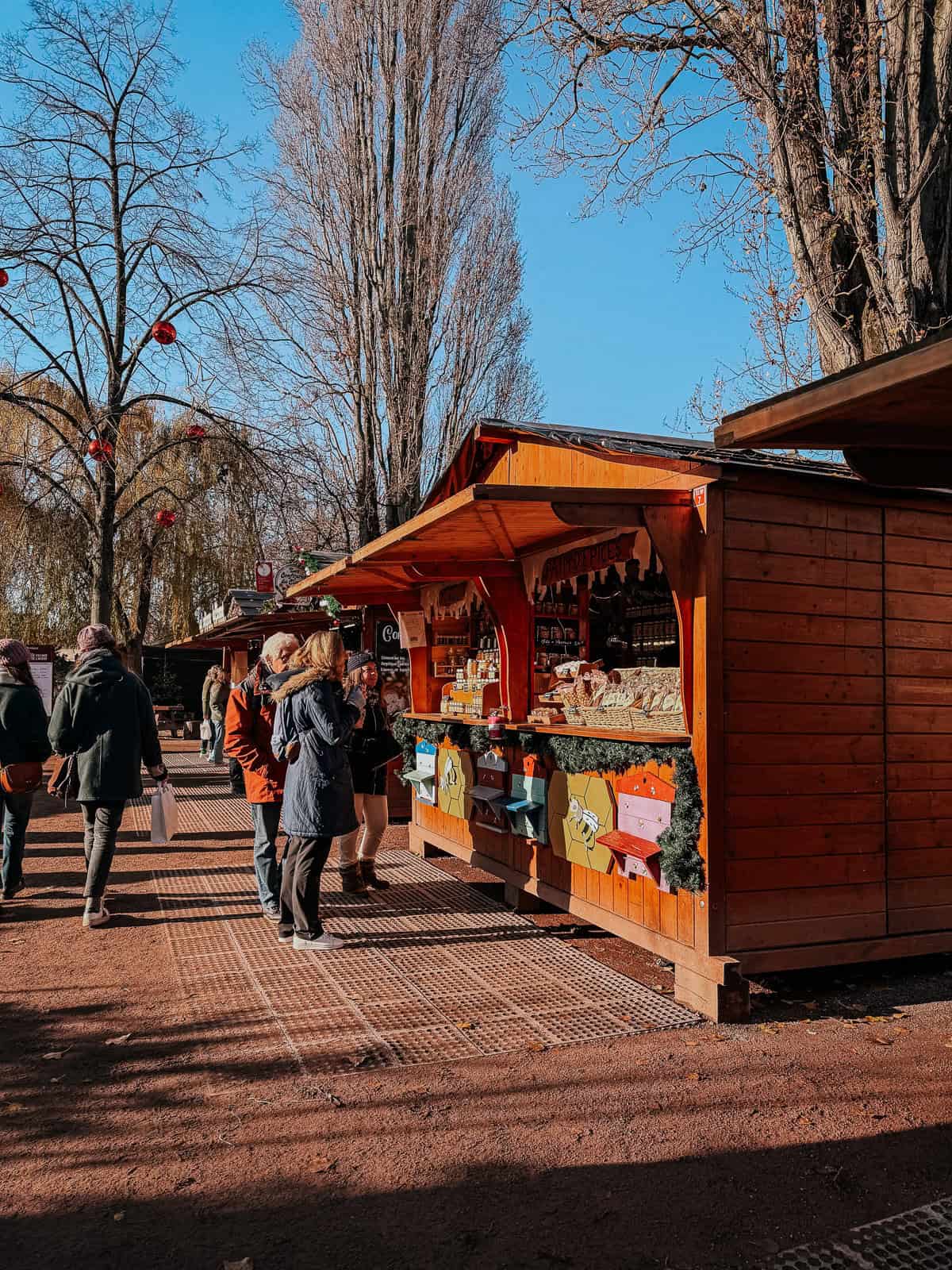 A sunny day at a Christmas market with shoppers walking past wooden stalls selling traditional goods under tall, leafless trees