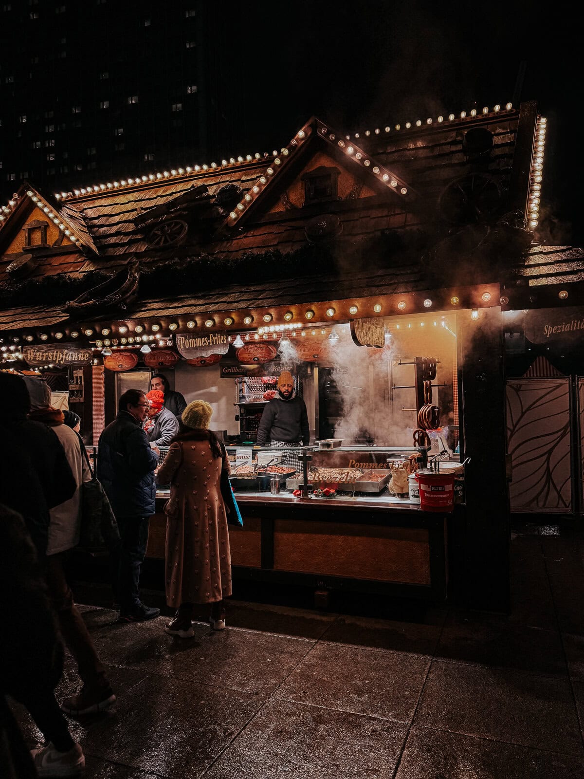 A bustling outdoor food stall at night, decorated with festive lights and emitting steam from cooking. People are gathered around, ordering and waiting for food, creating a lively winter market scene