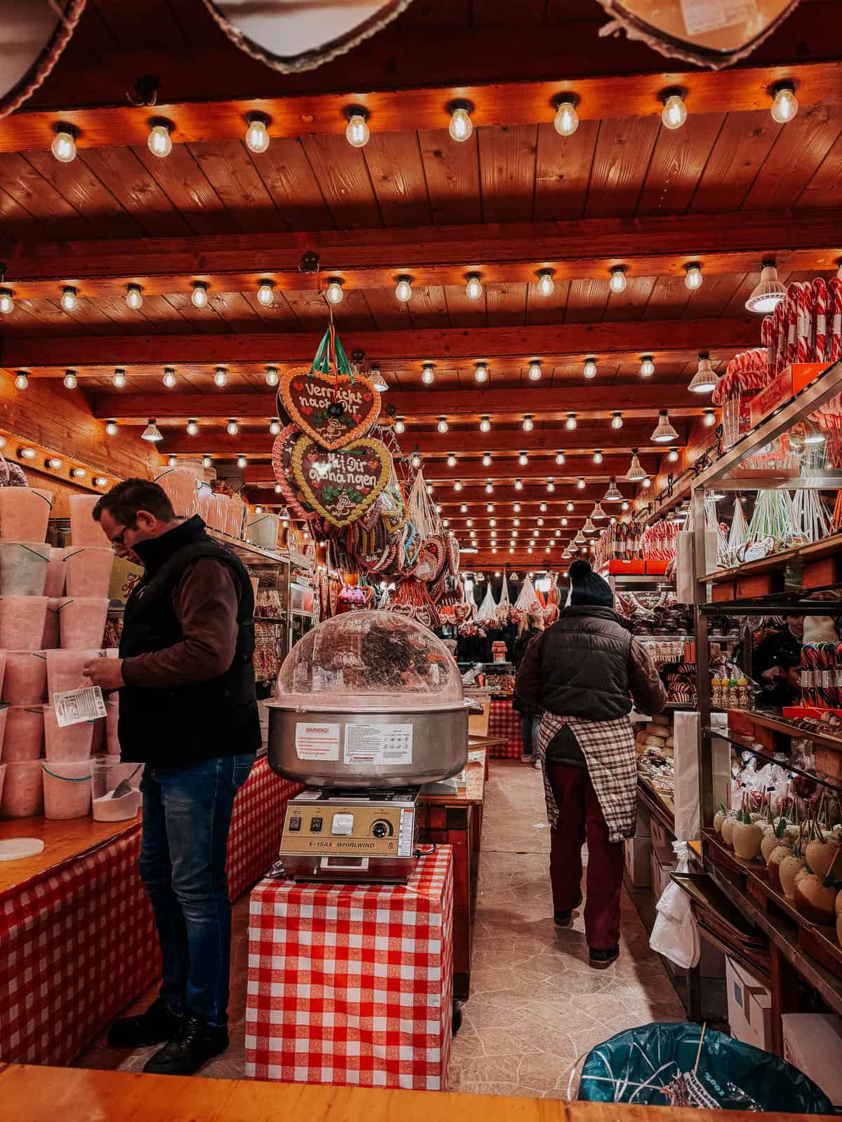 Inside a market stall lined with wood and warmly lit by overhead bulbs, showcasing an array of colorful candies and confections. Shoppers are seen browsing and purchasing, adding a busy and cheerful vibe to the setting