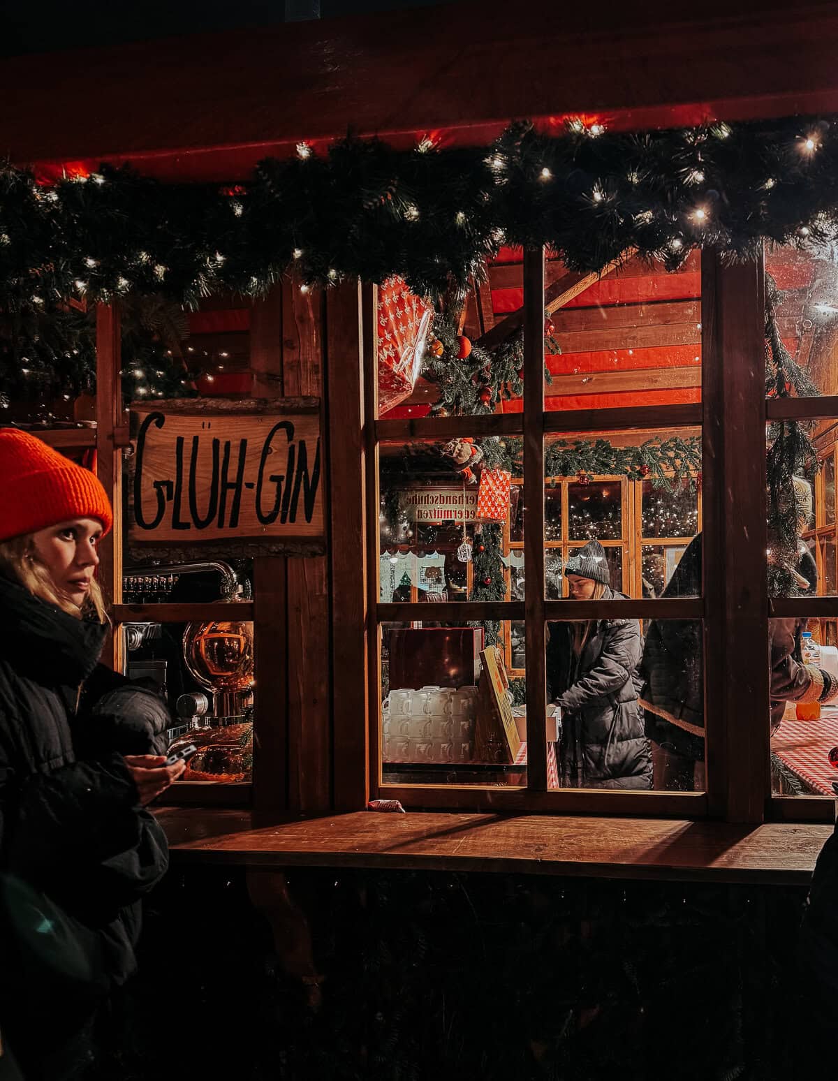 A detailed view of a market stall window decorated with festive garlands and a sign reading 'GLÜH-GIN.' Inside, patrons are visible interacting and browsing holiday-themed items, adding to the warm, festive mood.