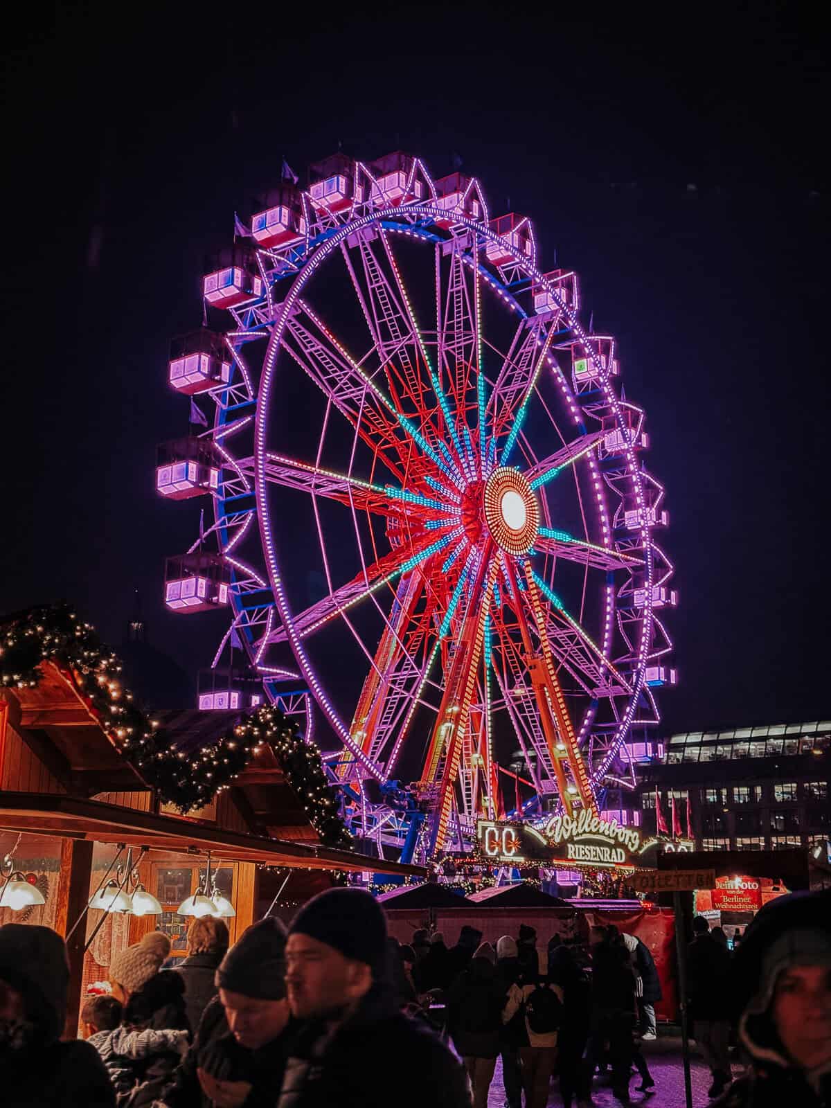 A vibrant and colorful Ferris wheel illuminated against the night sky at a bustling Christmas market. The wheel is surrounded by festive decorations and crowds of people, enhancing the festive atmosphere