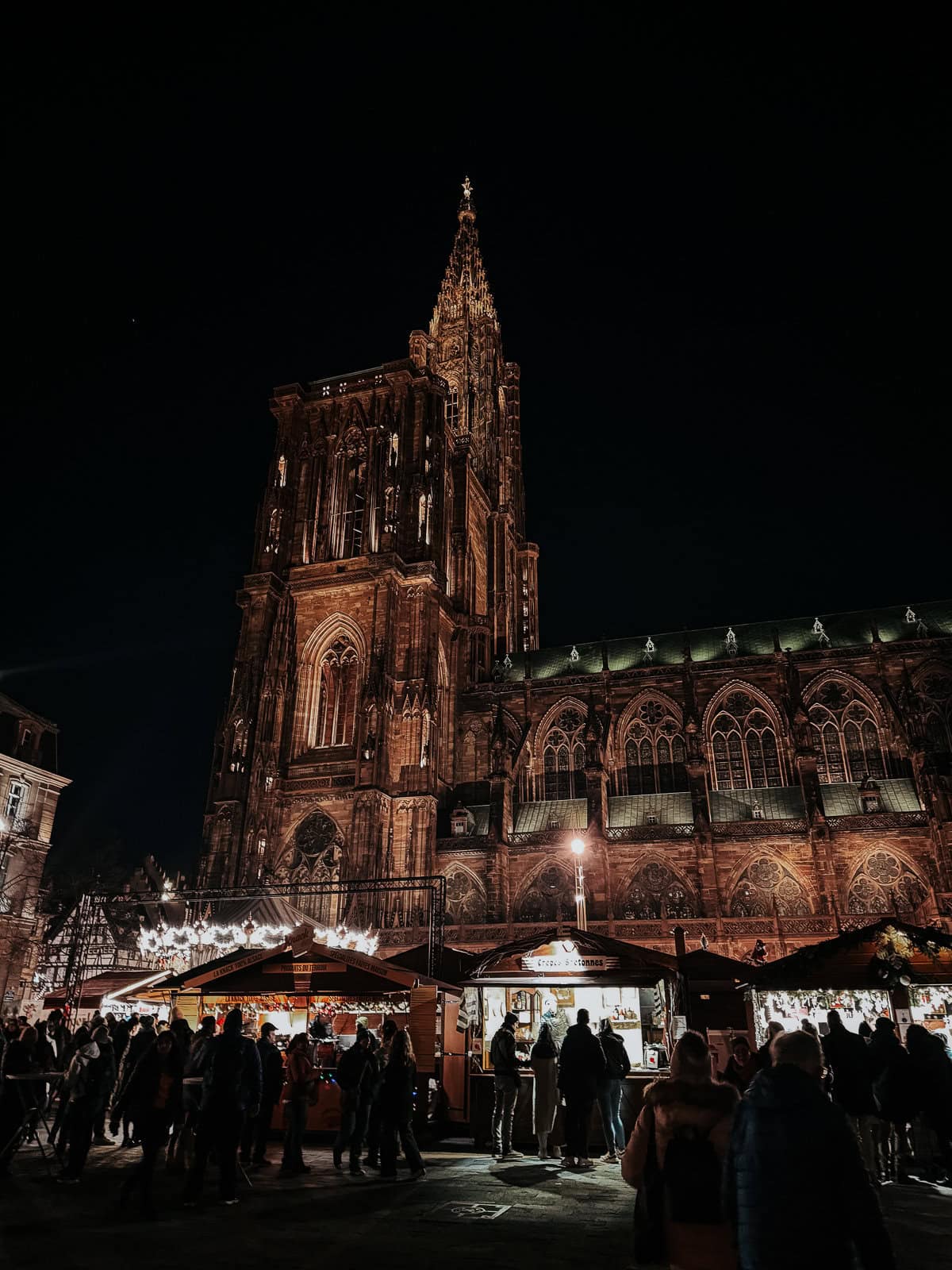A night view of an illuminated Gothic cathedral towering over a Christmas market bustling with people and decorated with white lights