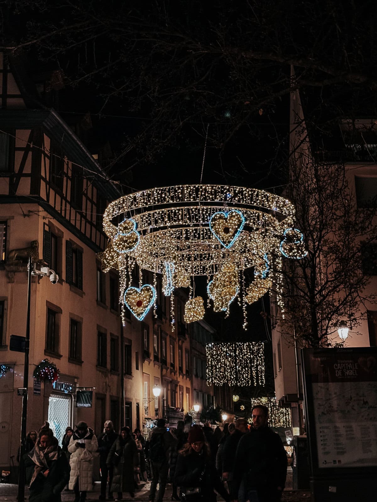 A festive street decoration featuring a large chandelier-like structure with glowing hearts and stars, set against a backdrop of traditional European buildings at night
