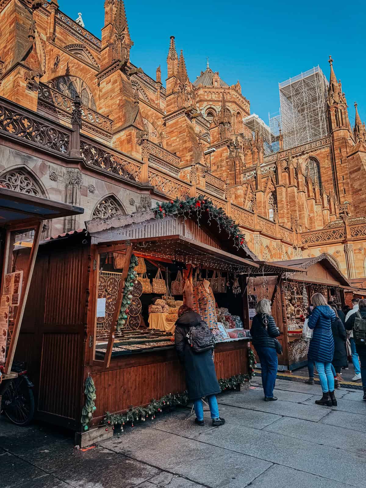 A majestic Gothic cathedral rising behind a Christmas market, with visitors gathered around various stalls under a clear blue sky.