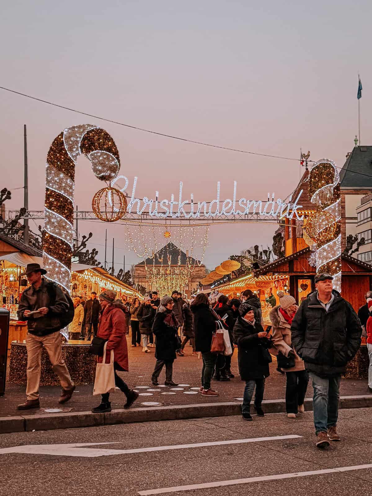A bustling Christmas market scene during twilight, showing rows of lit stalls under a sign reading 'Christkindelsmärik' with a large candy cane and festive decorations