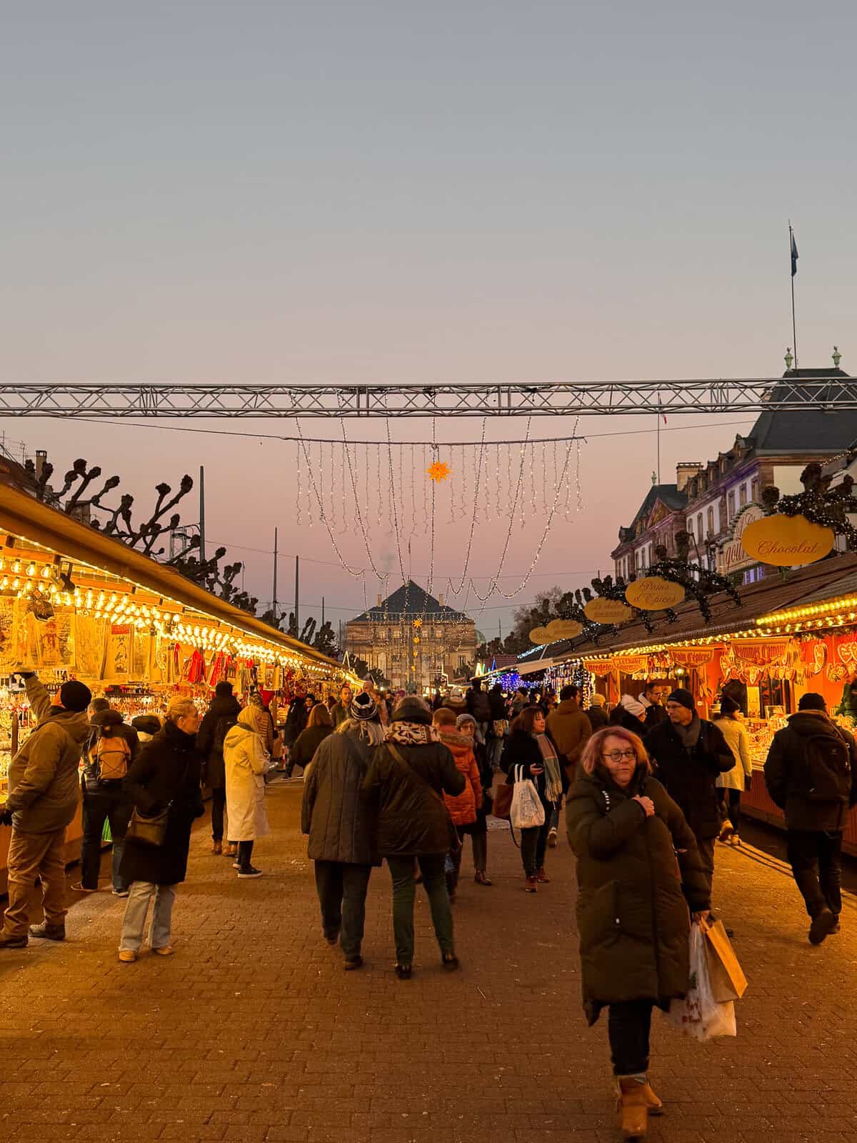 A vibrant Christmas market scene under a twilight sky, featuring an archway with the words 'Christkindelsmärik' illuminated by lights and surrounded by busy market sta