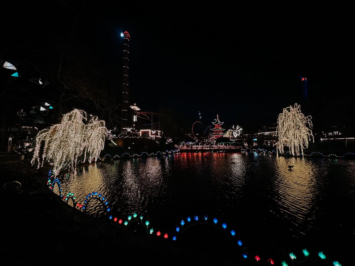 A festive view of Tivoli Gardens amusement park at night, illuminated with Christmas lights reflecting on a lake, showcasing rides and traditional architecture.