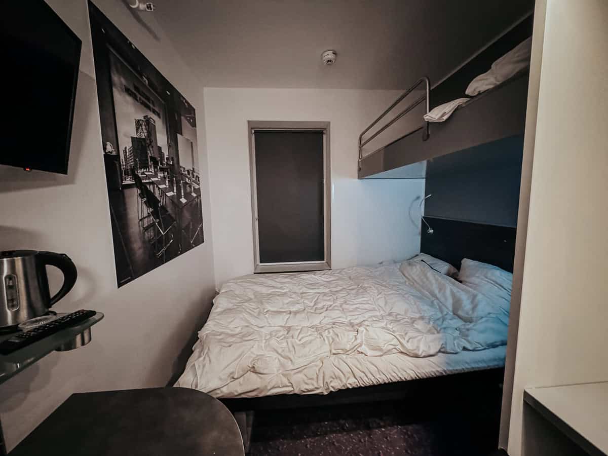 A compact, minimalist bedroom featuring a large bed with white bedding, a wall-mounted television, a kettle, and a large photograph of an industrial setting