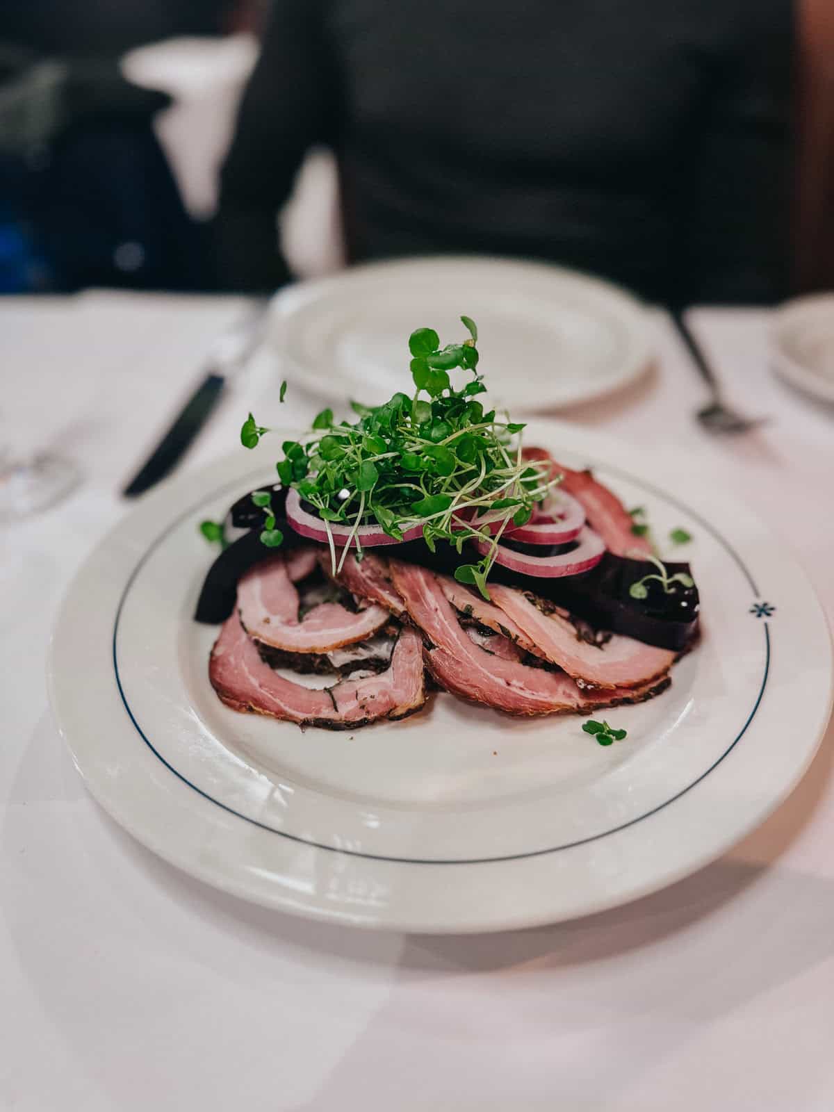 A well-presented plate of sliced roast duck garnished with green microgreens and red onion rings, served on a white plate with a blue-rimmed border in an elegant restaurant setting.