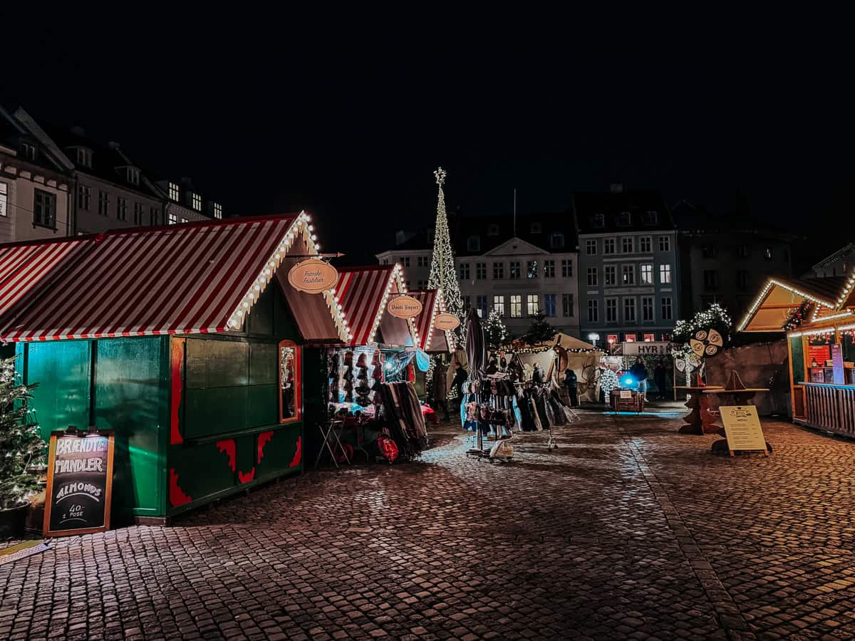 A vibrant evening scene at a Christmas market, showing various decorated stalls and a large crowd enjoying the festive atmosphere.