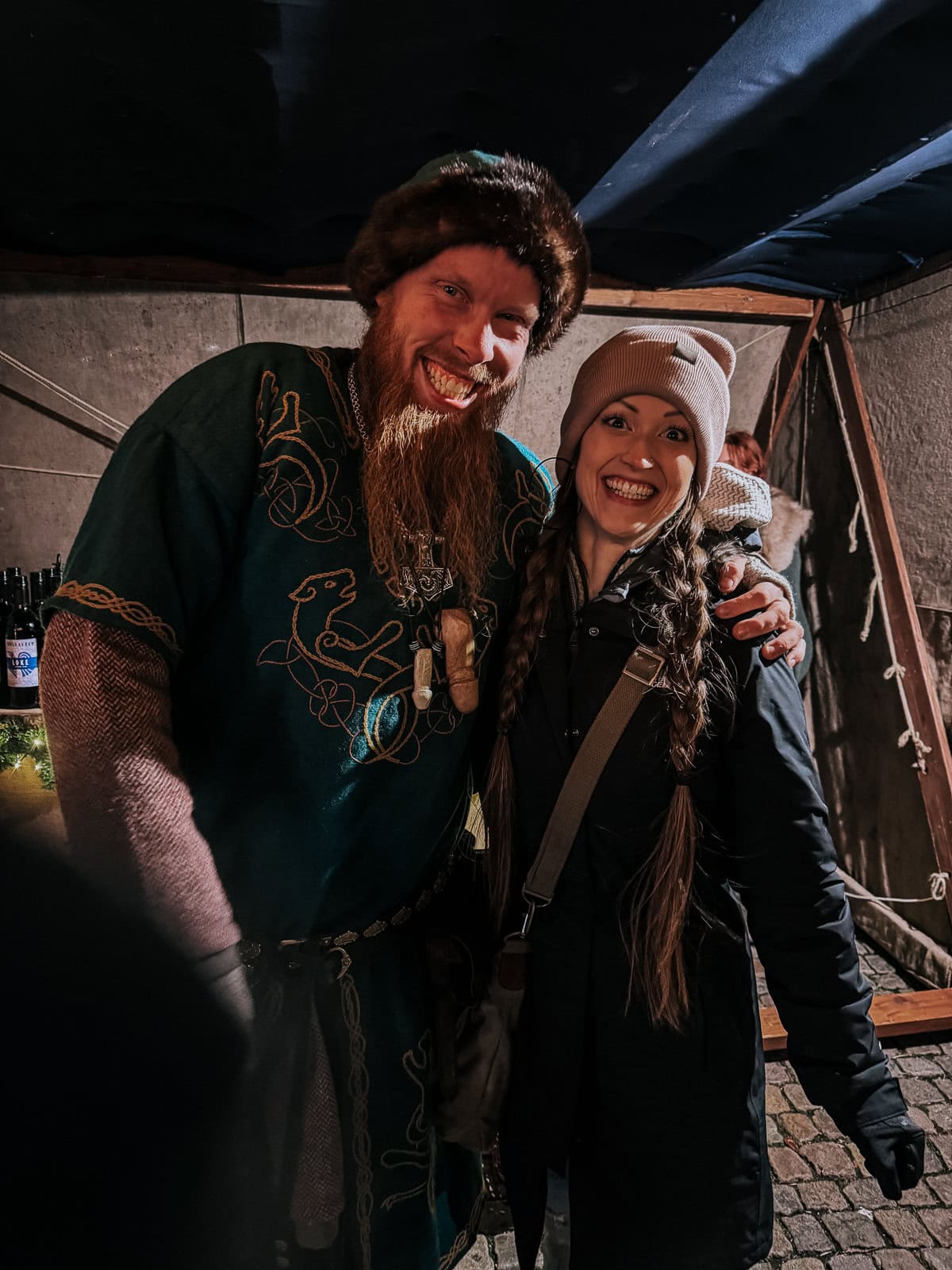 A man and a woman in Viking costumes smiling for a photo at a Christmas event, the man wearing a green tunic and fur hat, and the woman in a white beanie and black coat.