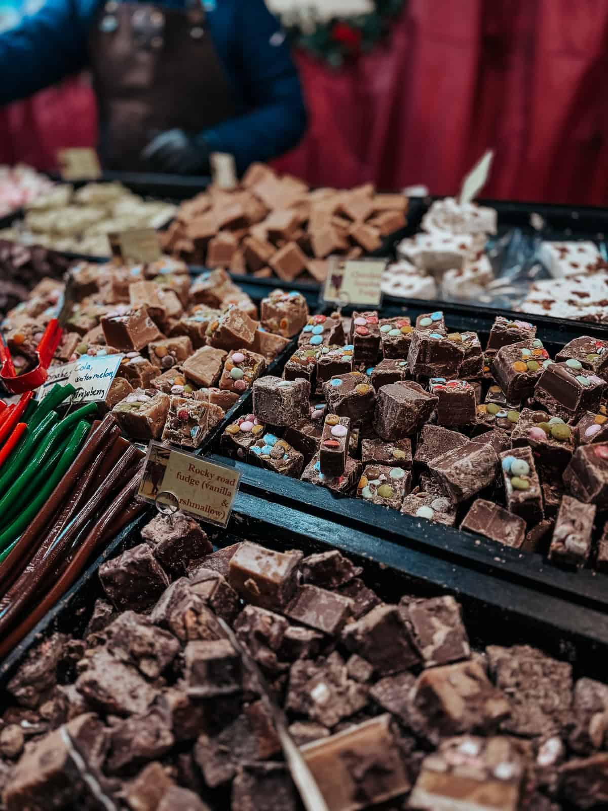 A close-up of various types of fudge, including chocolate, rocky road, and other candy-topped selections, displayed in a market stall with a vendor in the background.