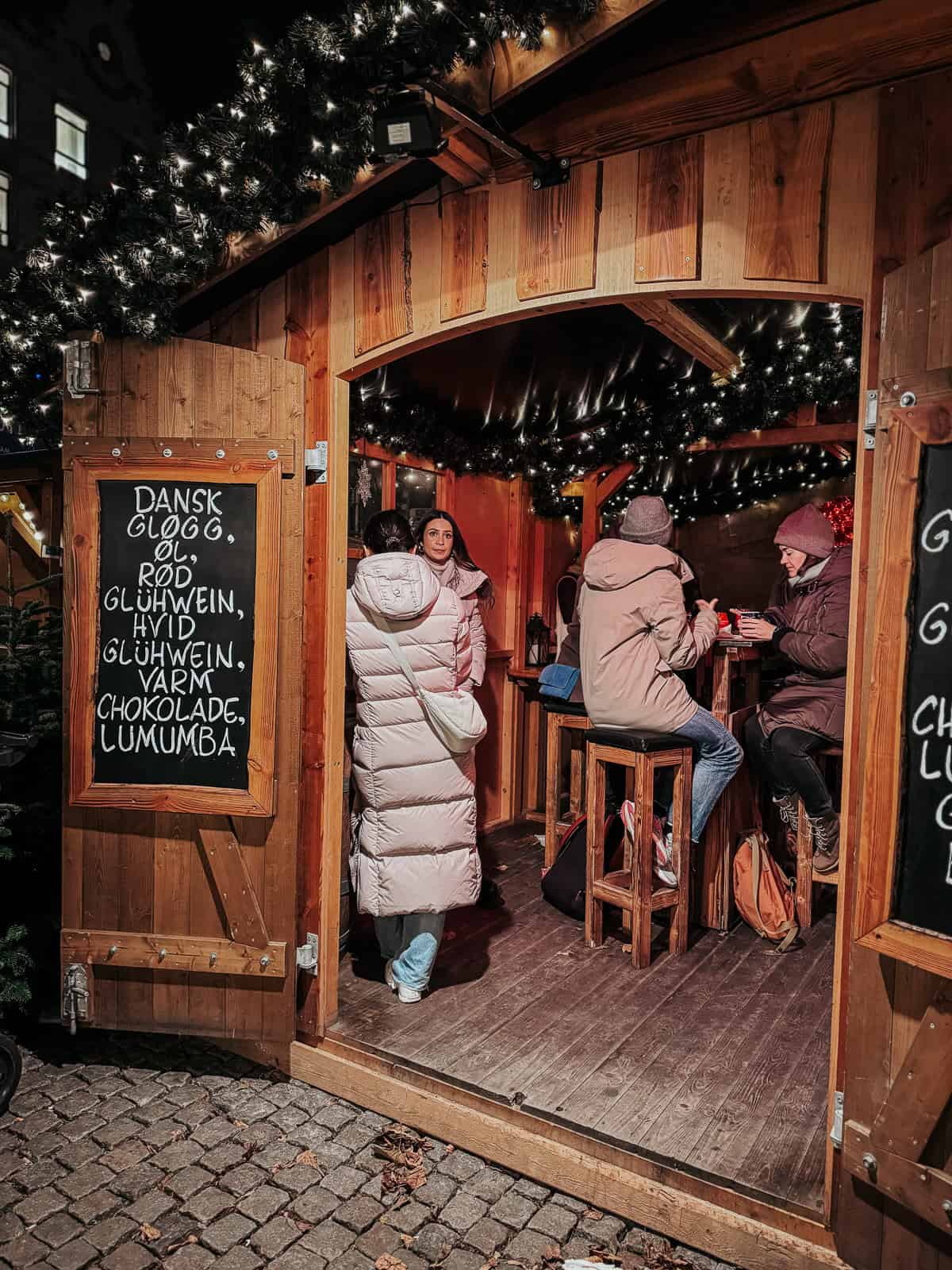 Patrons inside a cozy, wooden cabin at a Christmas market, illuminated by fairy lights and offering traditional winter drinks, as per the signage.