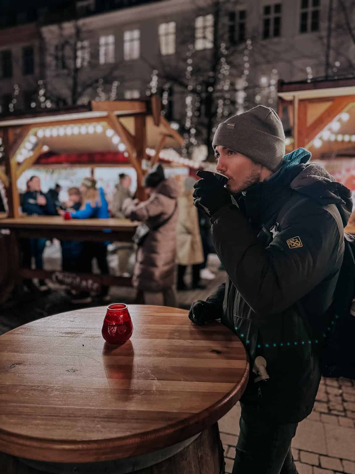 A man in winter attire drinks from a cup at a Christmas market, standing next to a wooden table with a red candle holder, with festive booths and sparkling lights in the background.