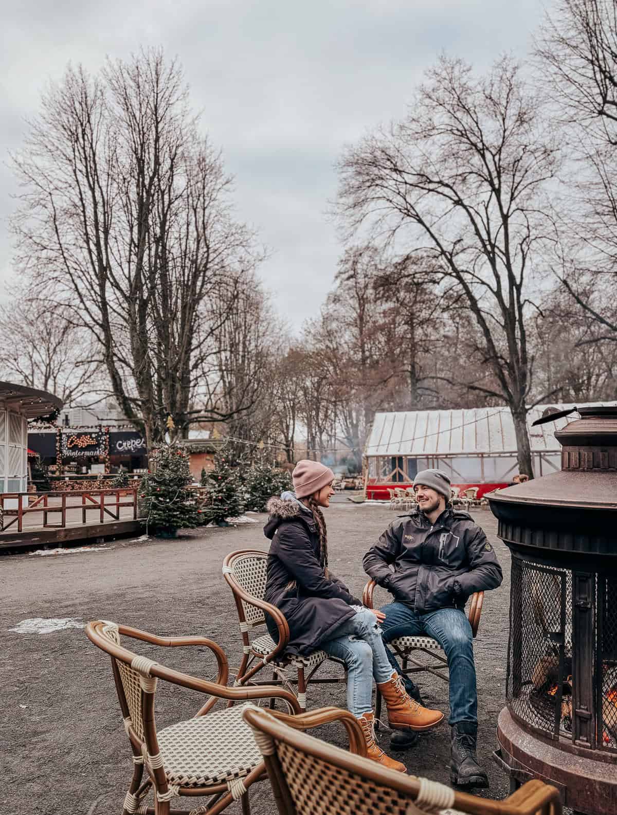 A cozy outdoor seating area with people warmly dressed, chatting near a lit stove. The setting is informal with rustic wooden chairs and festive decorations, nestled in a winter park.