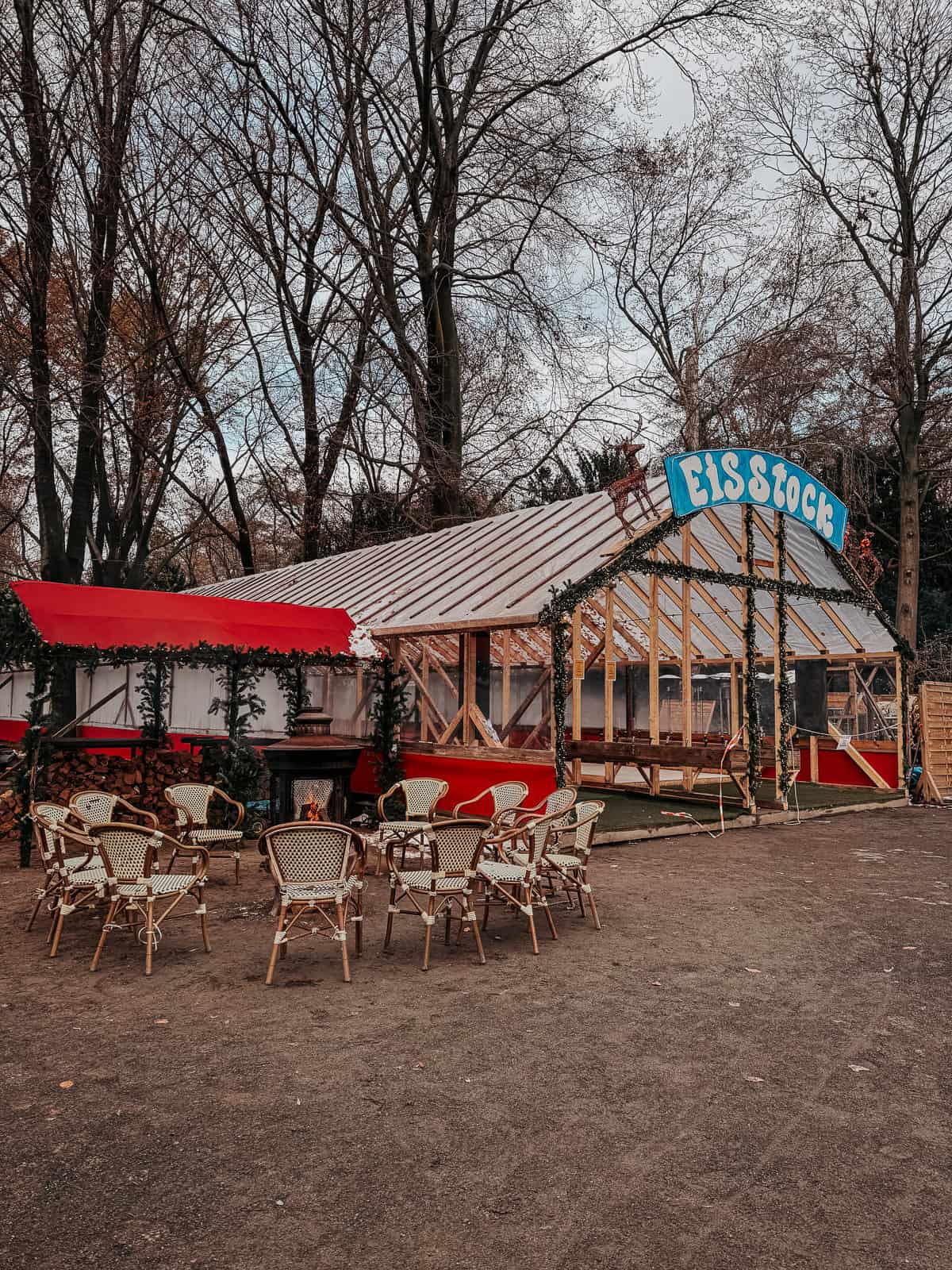 A quaint outdoor café with wicker chairs and red accents, set in a serene park setting during winter. The café appears closed, adding a peaceful contrast to the surrounding bare trees.