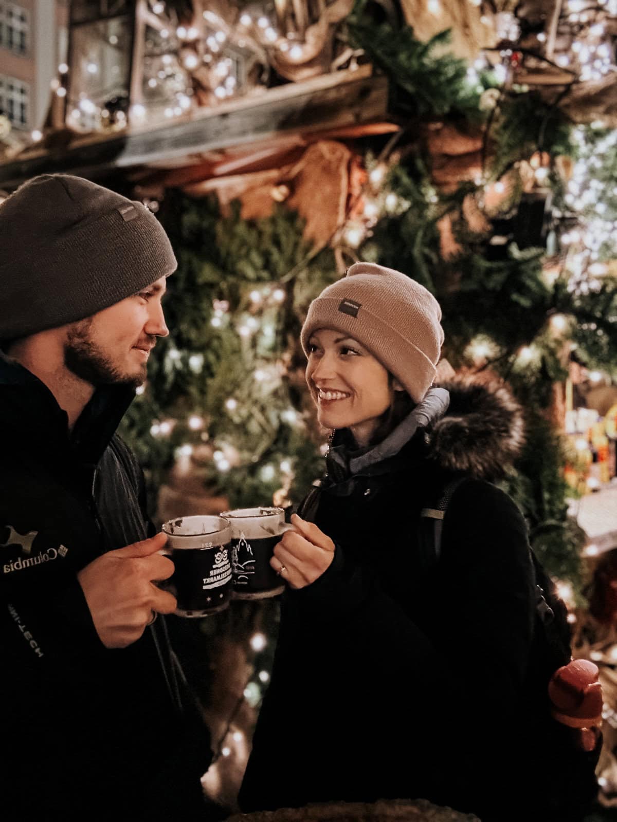 a couple stands in front a christmas tree doing a cheers with two clear glasses filled with a dark drink.