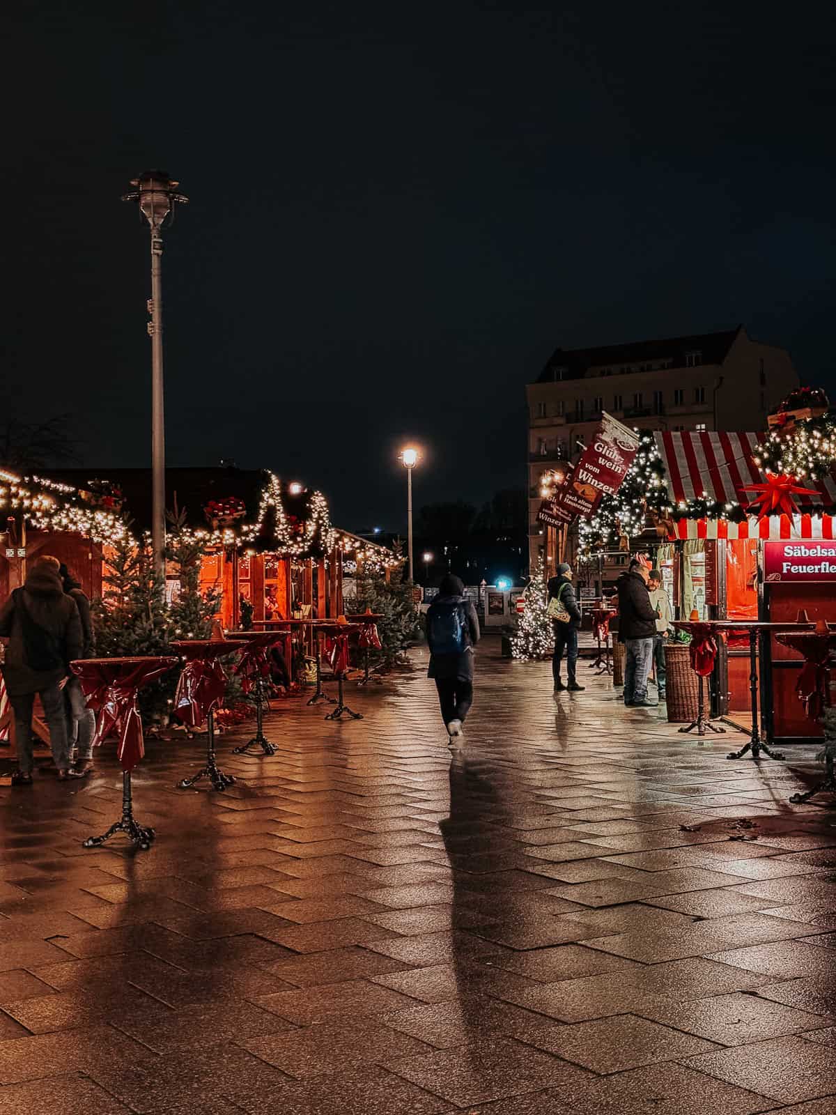 A well-lit pedestrian street adorned with festive decorations and Christmas lights. The scene shows empty café tables and people strolling along the pavement, contributing to a serene yet festive urban environment