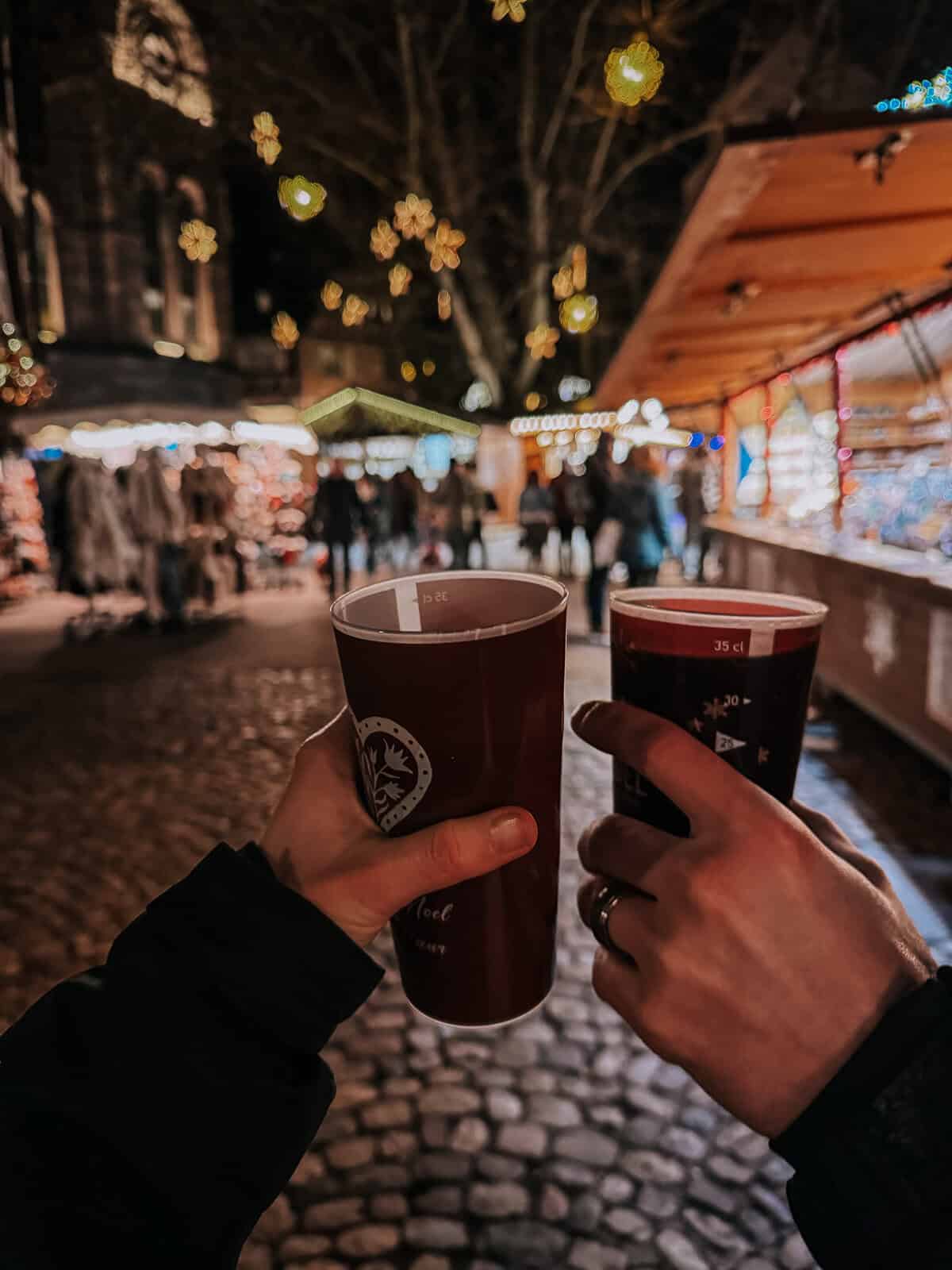 Two hands holding cups of mulled wine at a bustling Christmas market at night, with glowing festive lights and market stalls in the background.