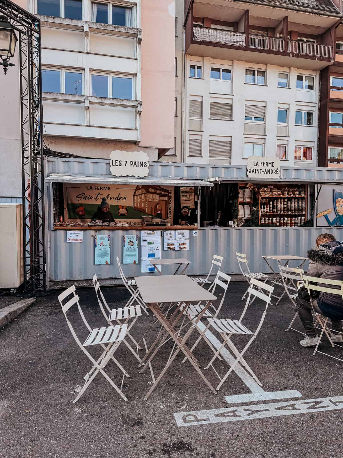A casual outdoor seating arrangement with metal chairs and tables in front of a small food stand, 'Les 7 Pains', set against an urban building backdrop.
