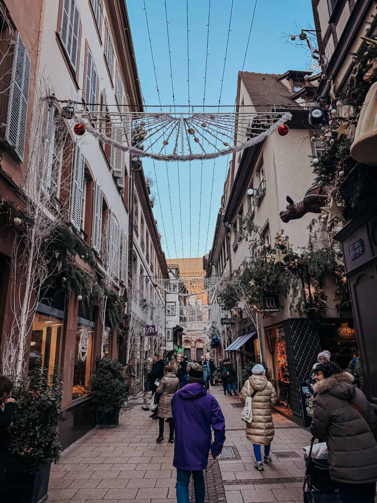 A narrow and bustling Christmas market street adorned with festive lights and decorations, flanked by traditional buildings and filled with holiday shoppers.