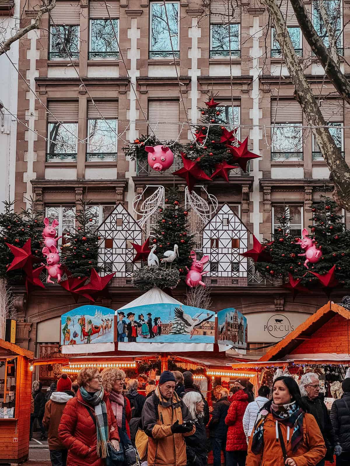 A building façade festively decorated with giant red stars and pink pig balloons, overhanging a Christmas market bustling with visitor