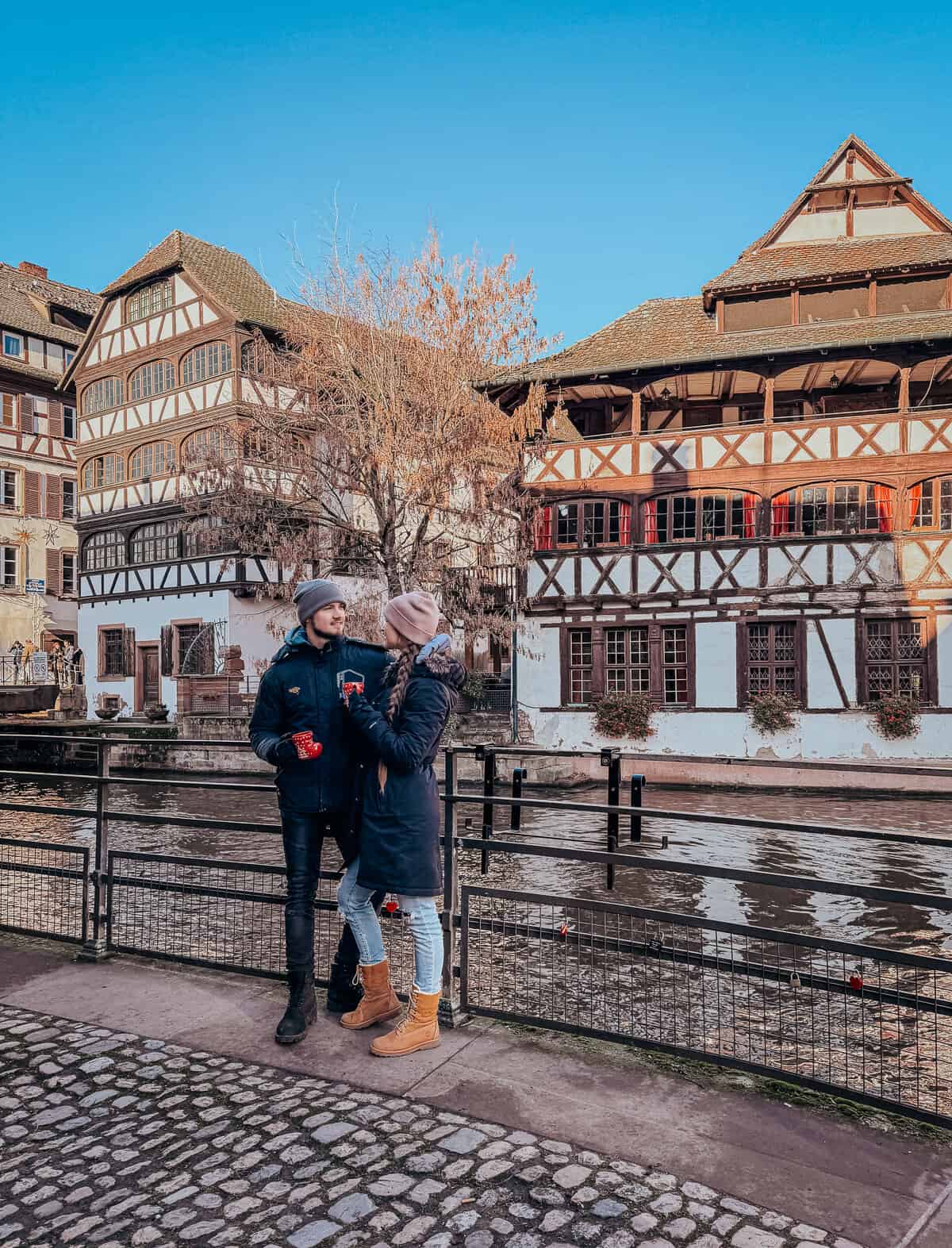 A couple enjoying mulled wine by a riverside, with traditional half-timbered houses in the background, evoking a romantic European winter scene.