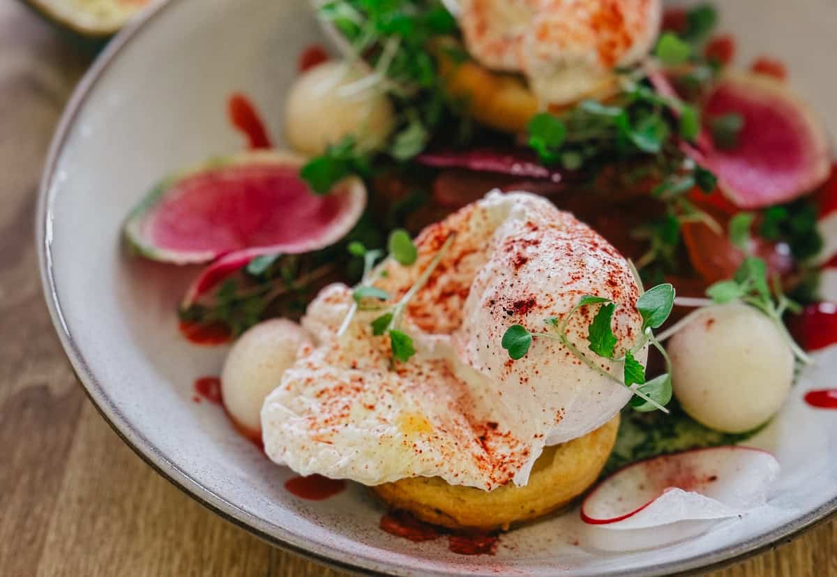 A close-up of a gourmet dish featuring poached eggs, garnished with microgreens and radish slices, served on a toasted base, with a colorful and artistic presentation.