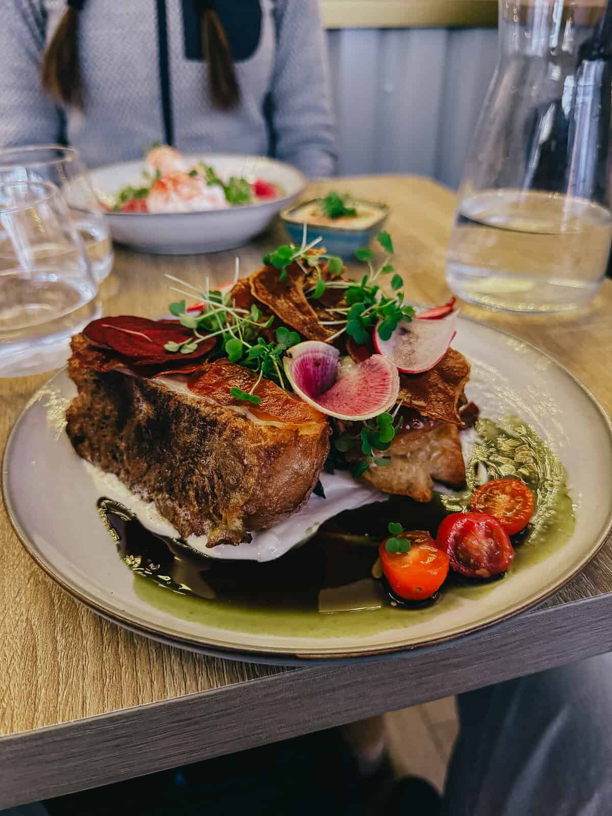 A plate of savory dish with thick bread, garnished with vibrant greens, cherry tomatoes, and radish slices, on a wooden table with a person in the background.