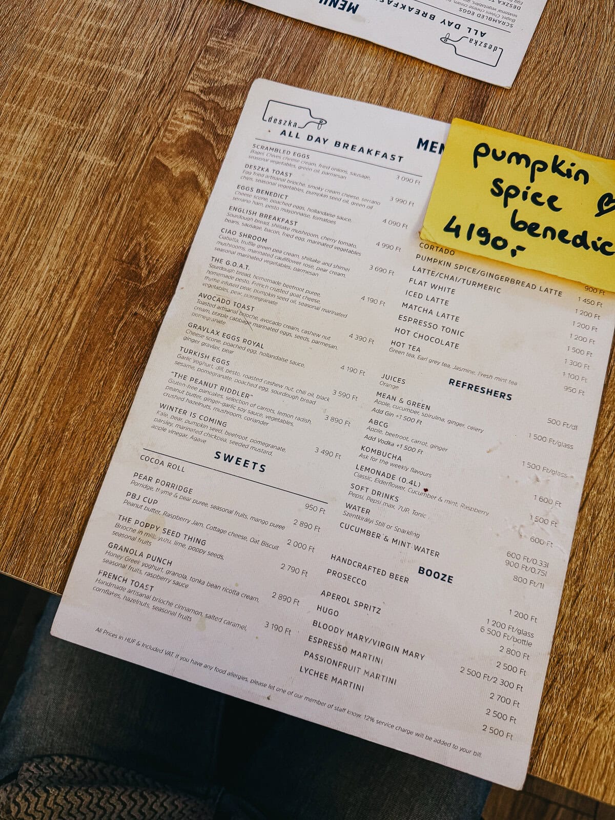 A cafe menu laid out on a wooden table, listing various breakfast items, beverages, and sweets, with a handwritten note highlighting 