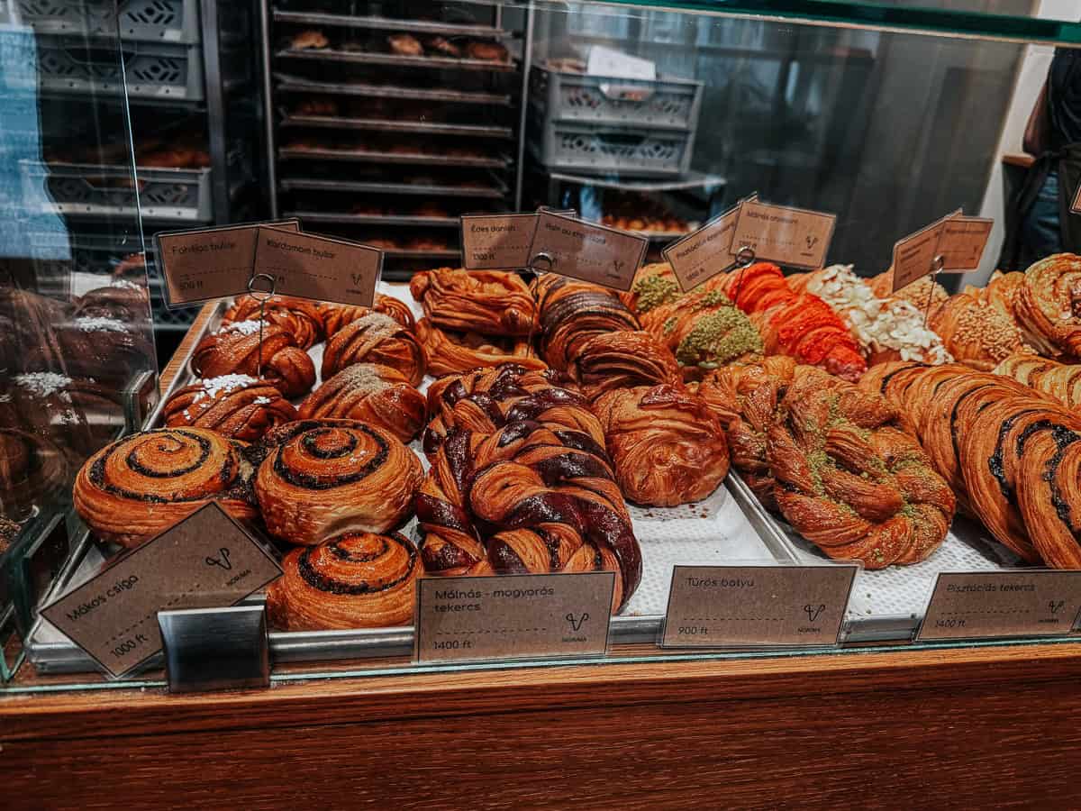 A bakery display case filled with a variety of pastries including cinnamon rolls, cardamom buns, and cheese-filled pastries, each labeled with small brown signs showing their names and prices in Hungarian forints.