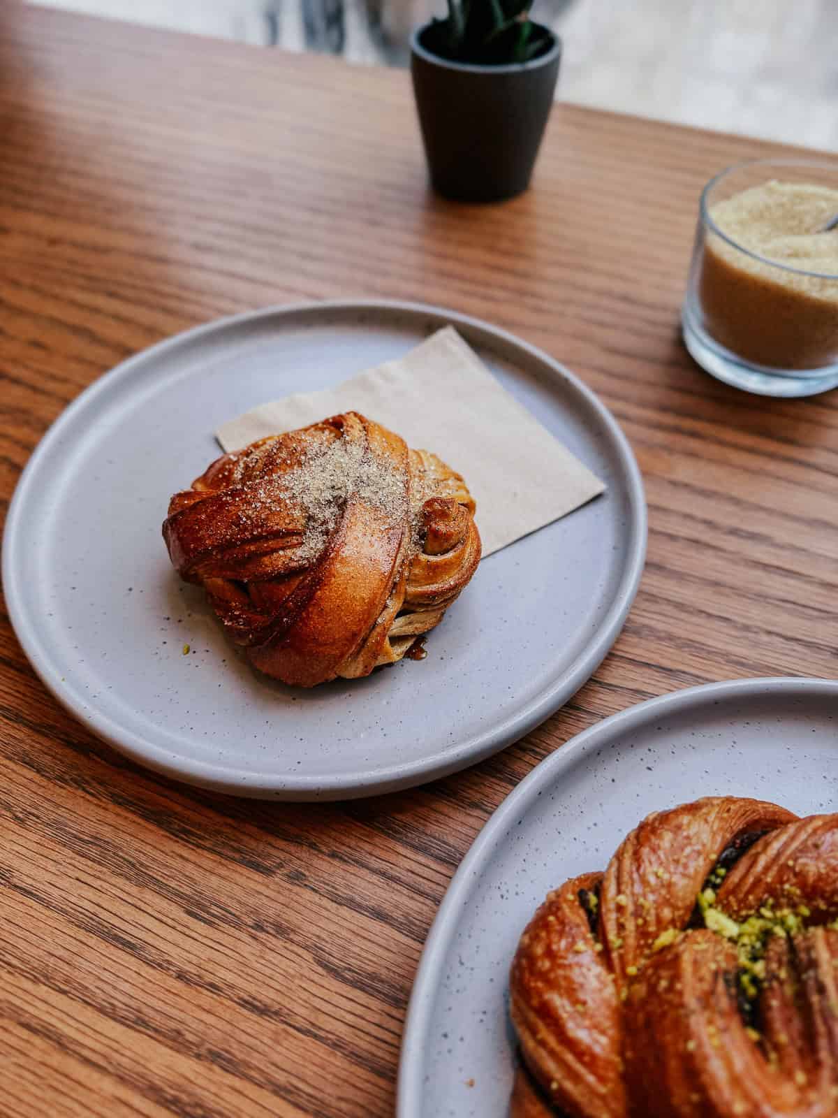 A cinnamon roll placed on a gray plate with a napkin, set on a wooden table with a small potted plant and a jar of brown sugar in the background.