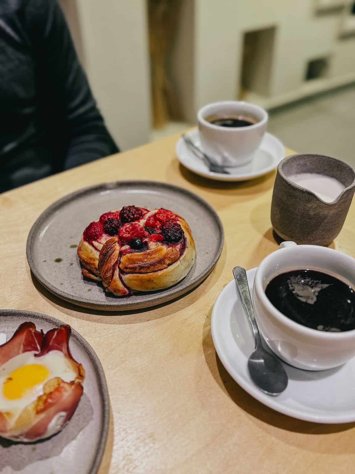 A table set with a cup of black coffee, a plate with a berry-topped pastry, and a ham-wrapped baked egg, with a person partially visible in the background.