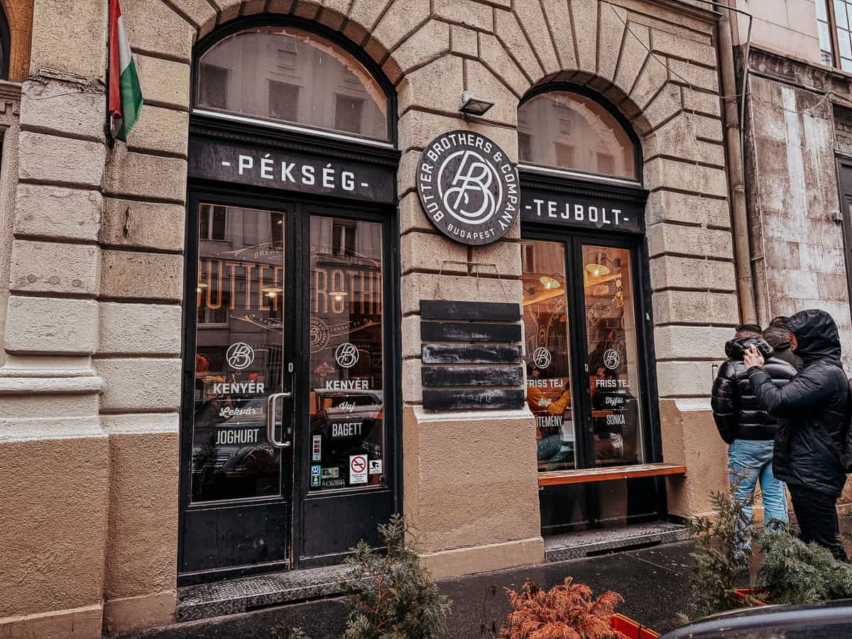 The exterior of Butter Brothers & Company, a bakery with large windows displaying the name and a Hungarian flag on the side, with two people standing in front.